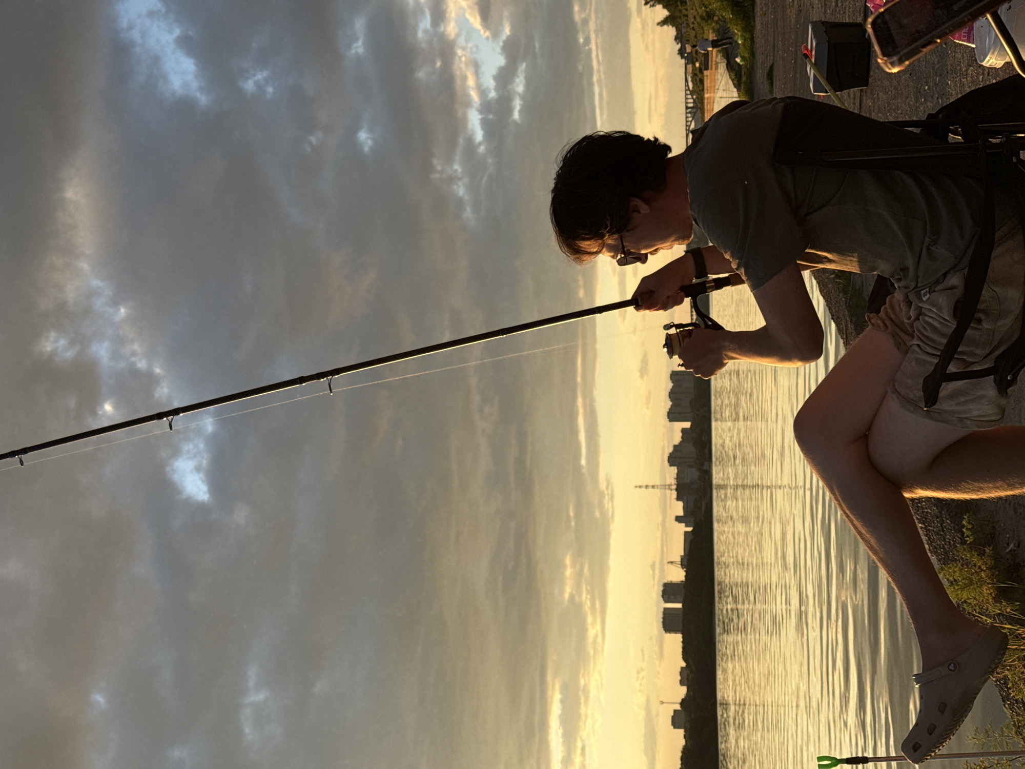 This media file captures a person fishing by a body of water during what appears to be sunset, casting long shadows and illuminating the sky with warm hues of orange and yellow. The person, dressed in a t-shirt and shorts, is seated and focused on their fishing rod, which extends across the frame. The water's surface reflects the dramatic sky, creating a shimmering effect. In the background, a cityscape with buildings and a distant tower is silhouetted against the bright horizon, suggesting an urban or semi-urban setting. The presence of a fishing chair and other equipment implies a dedicated fishing excursion. The overall mood is one of calm and concentration, with the natural beauty of the sunset adding a serene atmosphere to the scene. The image is oriented vertically.