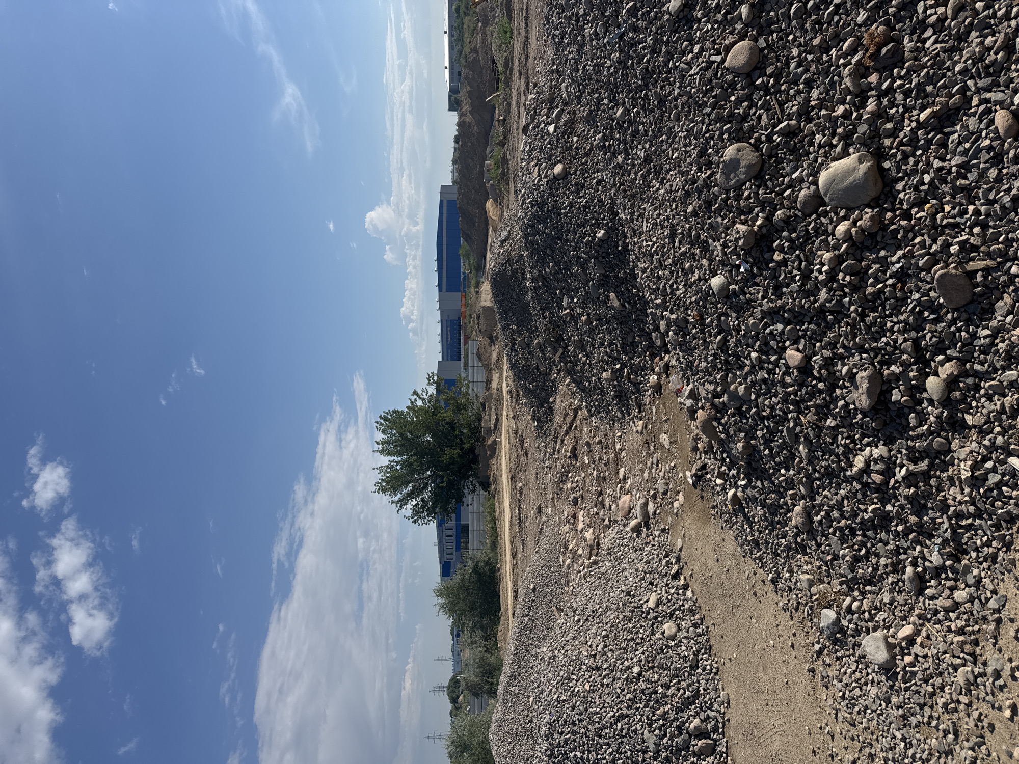 The image depicts an outdoor scene in Köközek, Kazakhstan, featuring a construction or material staging area under a bright, partly cloudy blue sky. In the foreground, several large piles of gravel and smaller rocks dominate the view. A dirt path or track runs between two of these piles. In the midground, more piles of earth and gravel are visible, along with scattered vegetation and some trees. Behind these, a large, modern building with blue and grey elements is situated, suggesting an industrial or commercial facility. In the distance, power transmission towers are visible on the horizon. The overall impression is one of a functional, undeveloped or under-development area within a semi-urban or industrial zone. There are no people or animals present in the image.
