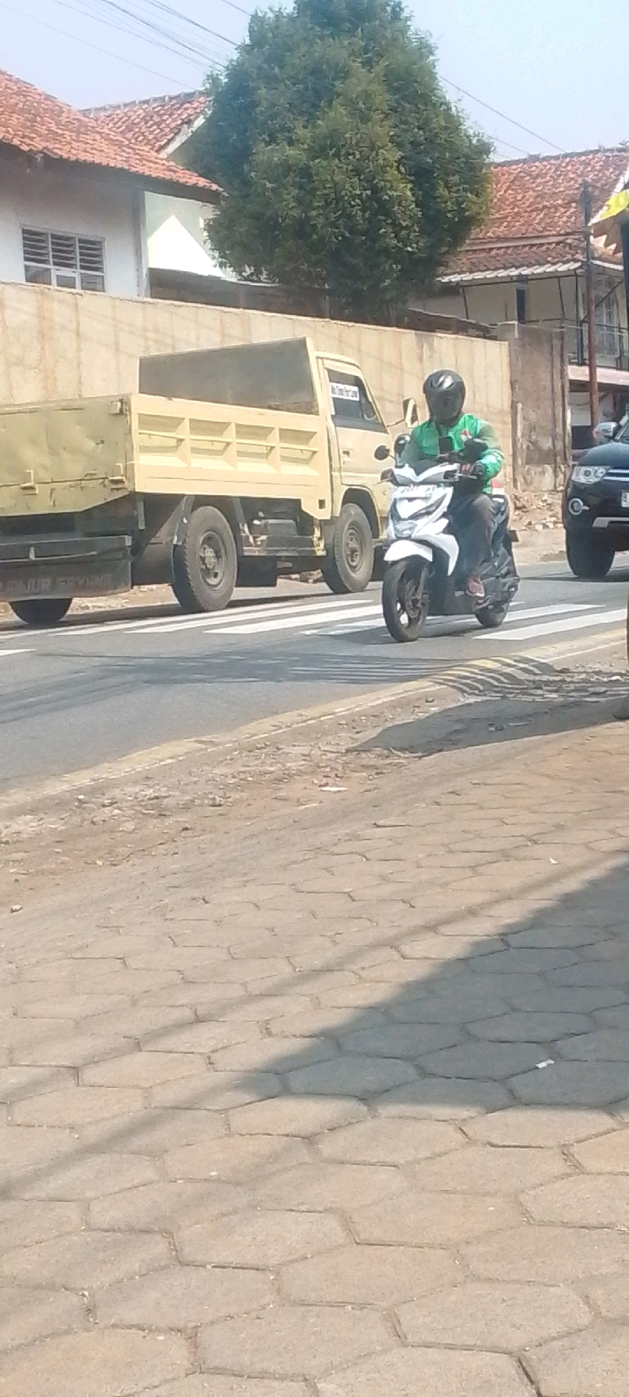 A man wearing a green jacket and helmet is riding a white motorcycle across a pedestrian crossing on a street in Sirnagalih, Indonesia. The man is in the middle of the crossing, and the motorcycle is angled slightly towards the right. In the background, a light yellow dump truck is stopped at the crossing, and to the right of the motorcycle, a black SUV is partially visible. The street is paved with asphalt, and the pedestrian crossing is marked with white stripes. Buildings with red tile roofs are visible in the background, along with a large green tree. The sky is clear and blue. The foreground shows a paved walkway with hexagonal stones.