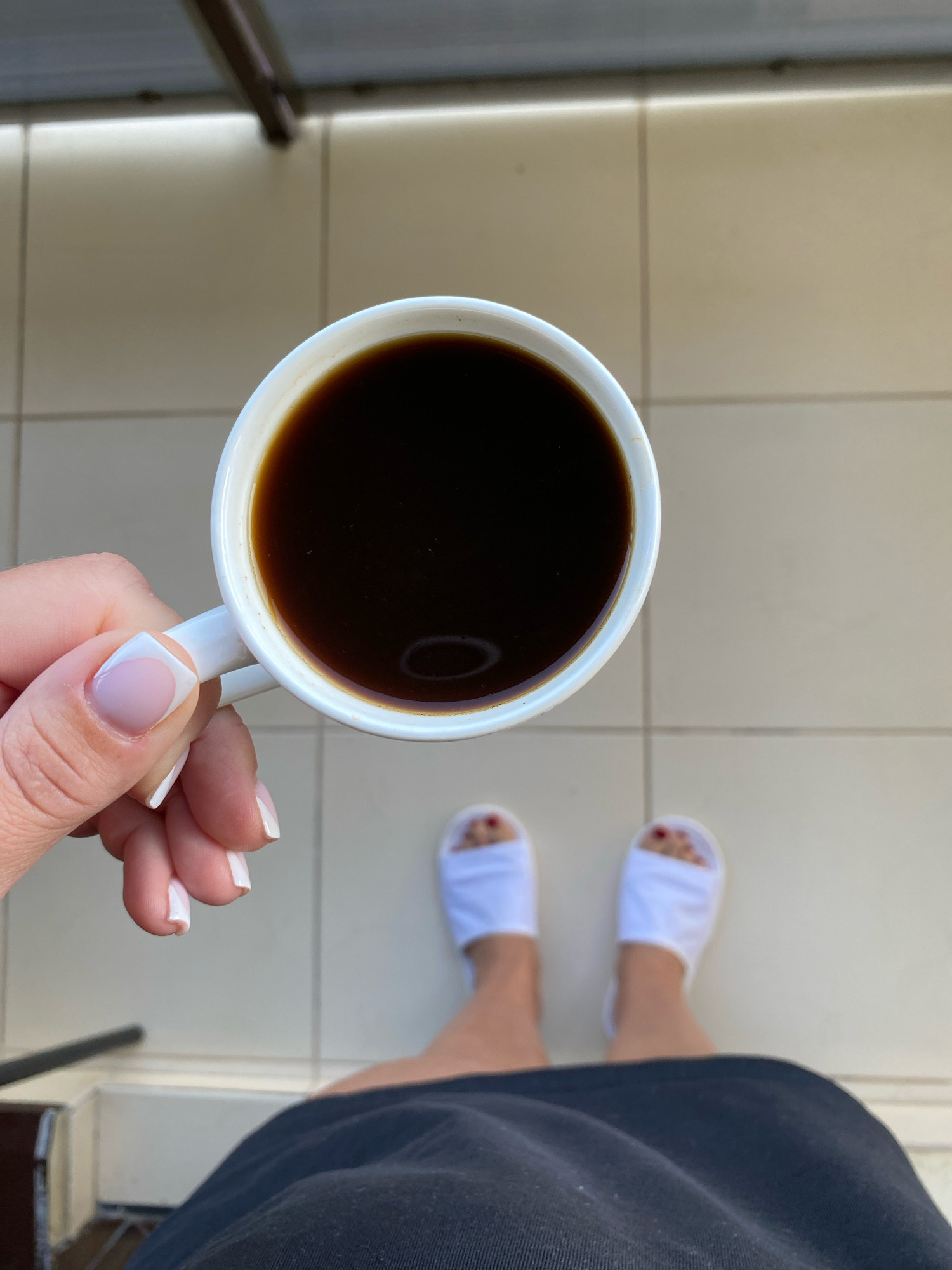 A first-person perspective shot shows a hand holding a white mug filled with dark coffee. The hand has French manicured nails. The person is wearing white slippers, and their feet are on a light-colored tiled surface. Part of a dark garment is visible at the bottom of the frame. The background consists of light-colored tiles and a railing. The setting appears to be an outdoor balcony or patio, potentially in Nebug, Russia.