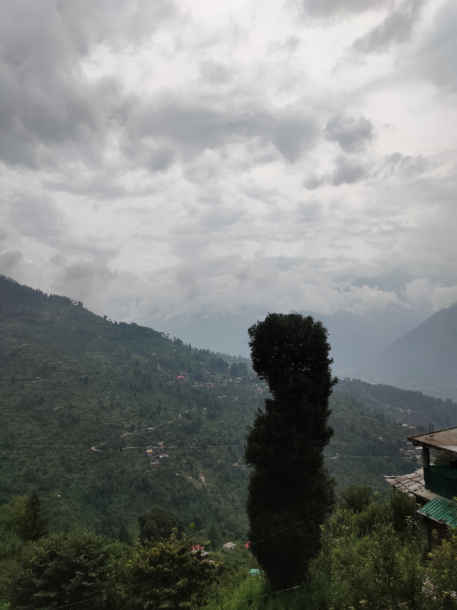 The image depicts a cloudy, overcast day in a mountainous region, likely in or near Raman, India. The landscape is characterized by steep, verdant hills covered in dense forestation. Scattered across the hillsides are small clusters of houses and buildings, with some winding roads visible. In the foreground, a tall, dark green tree stands prominently. To the right, part of a building with a sloped, green roof and a flat concrete roof is visible, suggesting human habitation within the natural environment. The overall atmosphere is serene and somewhat muted due to the cloudy sky, creating a sense of depth and distance within the scene.