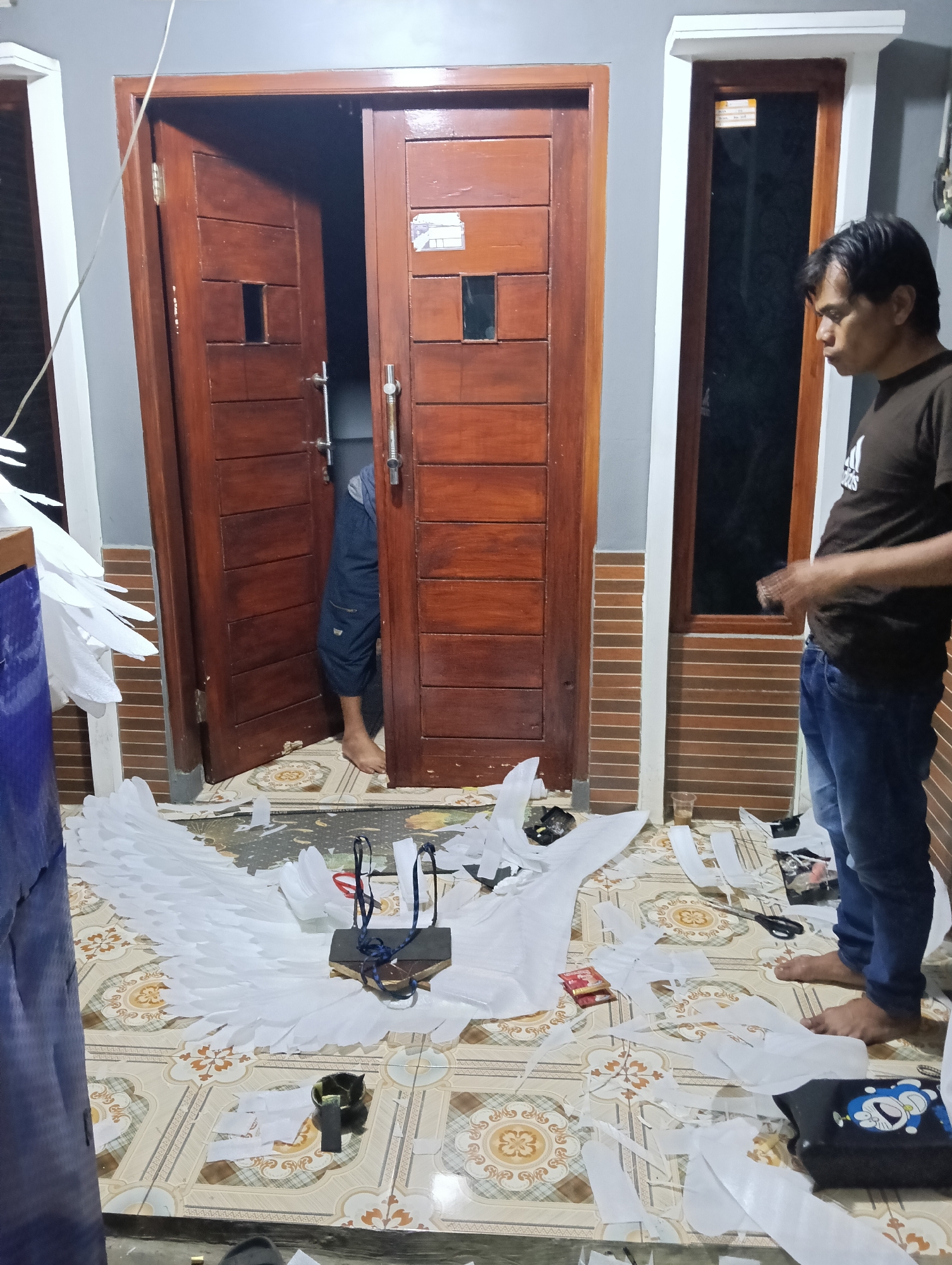 In Tasikmalaya, Indonesia, a person is in the process of constructing large white wings, possibly for a costume. The wings, made of white material and feathers, are spread out on the floor. Scattered around are scraps of the white material, scissors, and other crafting supplies. The person, wearing a brown t-shirt and blue jeans, is standing and observing the work. A wooden door stands open in the background, and a window with wooden trim is visible on the right. The floor is tiled with a patterned design.