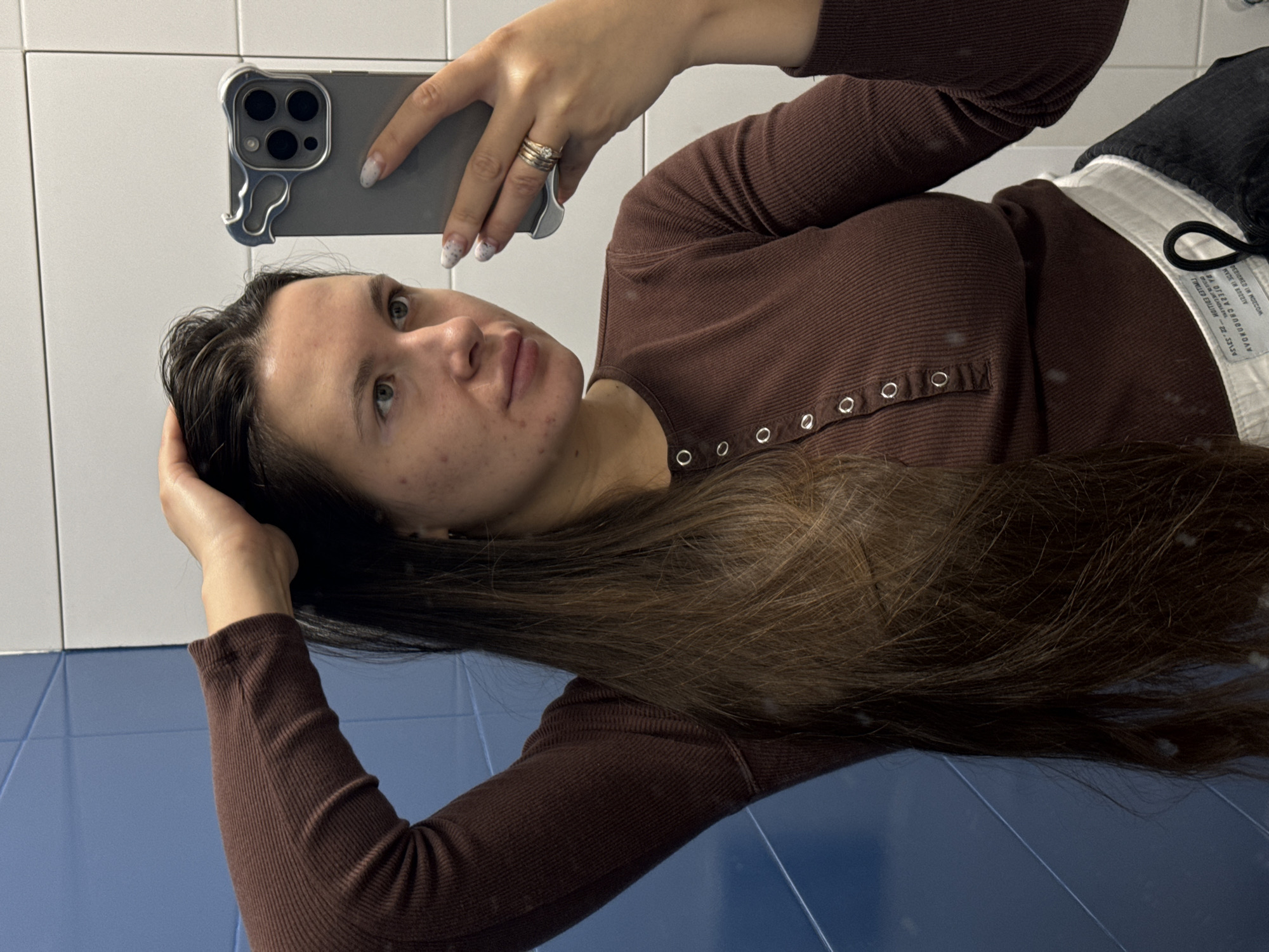 A woman takes a selfie in a bathroom. She holds a gray smartphone with a unique case, displaying patterned fingernails and rings on her fingers. She is wearing a brown long-sleeved shirt with snap buttons down the front. A portion of her pants is visible, showing a black fabric and a white waistband with text. The bathroom features white tiled walls above a blue tile floor or paneling. There is no indication of the image being taken in Odintsovo, Russia.