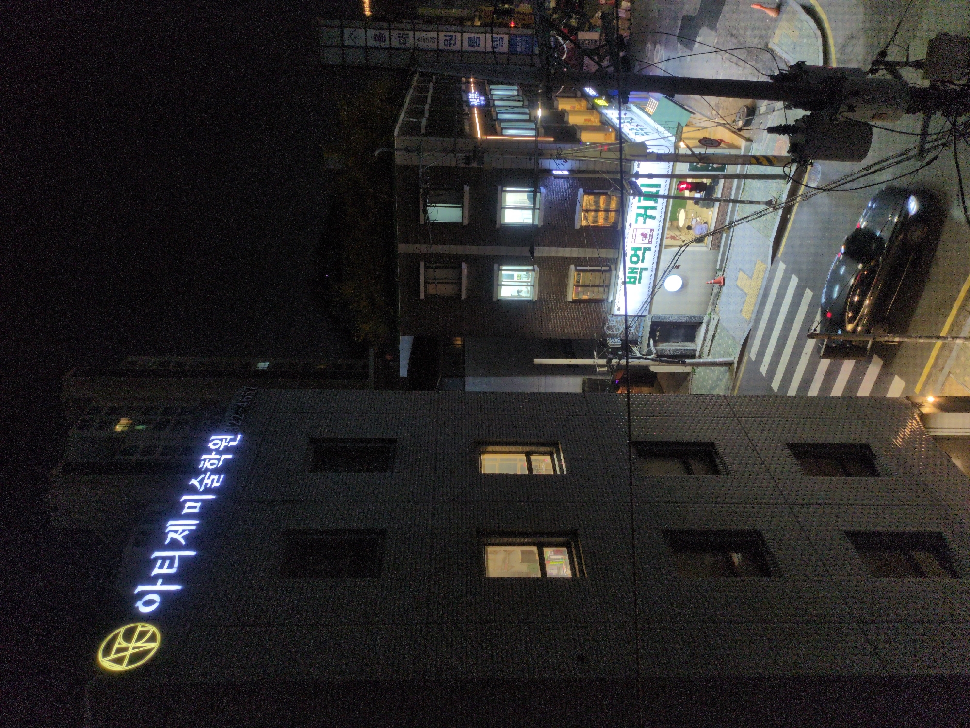 At night, the camera is positioned at street level in an unknown city in Unknown, looking toward a street intersection. A dark grey, multi-story building dominates the foreground, its façade punctuated by windows, some of which are illuminated. Vertically displayed in bright white neon lettering on the building is Korean text. Further away, another multi-story building is visible with illuminated windows and storefronts. A traffic light above a crosswalk is red. A black car is shown on the right side of the road near a power line.