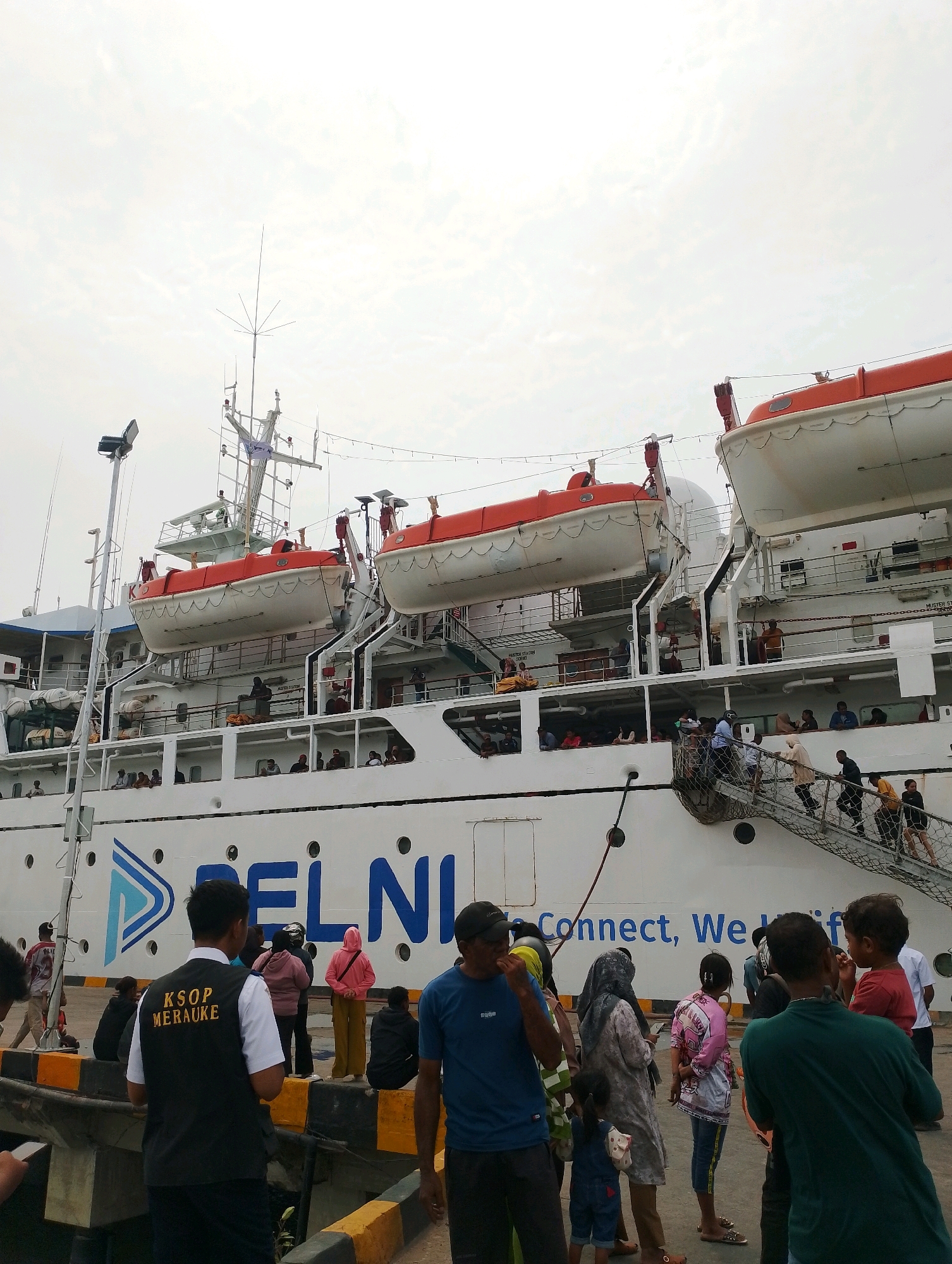 A large white passenger ship, identified by the logo "PELNI" and text reading "Connect, We Unite," is docked at a harbor in Merauke, Indonesia. The ship has multiple decks with lifeboats visible. A crowd of people, including men, women, and children, are gathered on the dock and boarding the ship via a ramp. Some individuals appear to be staff, one of whom is wearing a vest with "KSOP MERAUKE" written on it. The environment is overcast, suggesting a cloudy day. The scene depicts a port operation with passengers preparing to board a vessel.