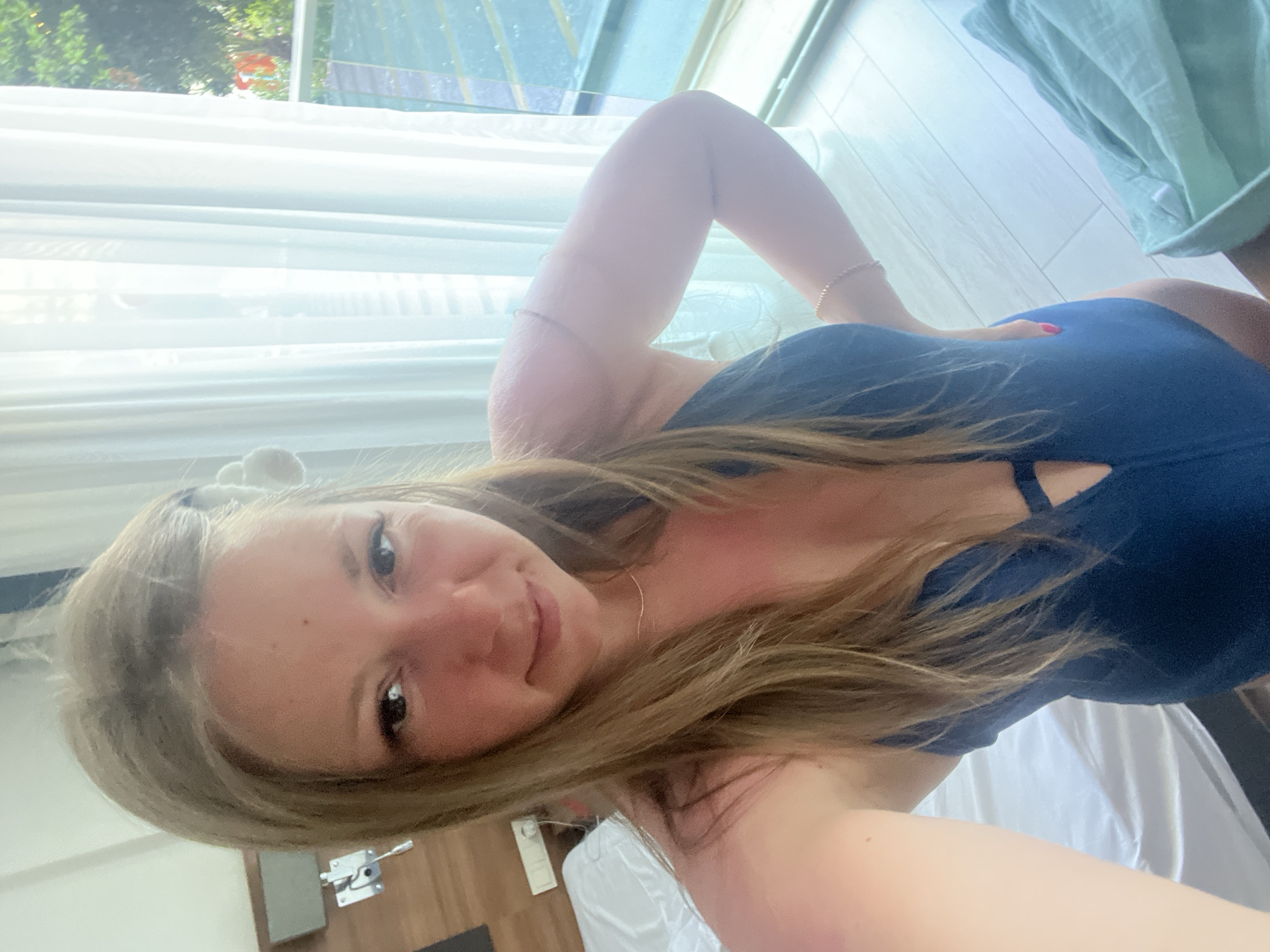 The image features a young woman lying on a bed, with her body positioned diagonally across the frame. She is wearing a dark blue one-piece swimsuit. Her face is turned towards the camera, and she is smiling. Her long, blonde hair is spread out around her head and shoulders. Her left arm is bent and resting on her head, while her right hand is placed on her swimsuit. A delicate bracelet is visible on her left wrist, and another on her right ankle.
The setting appears to be an indoor space with large windows that allow natural light to filter in, illuminating the room. White curtains hang by the windows, and sheer white fabric, possibly a bedspread or sheet, covers a surface beneath her. A wooden floor is visible in the background, and a light green fabric, possibly a towel or blanket, is draped over something to the right of the frame. Outside the window, greenery and a glimpse of what appears to be a swimming pool or an outdoor area can be seen, suggesting a hotel or resort setting. The lighting suggests it is daytime. The overall mood is relaxed and cheerful. There is no visible text in the image.