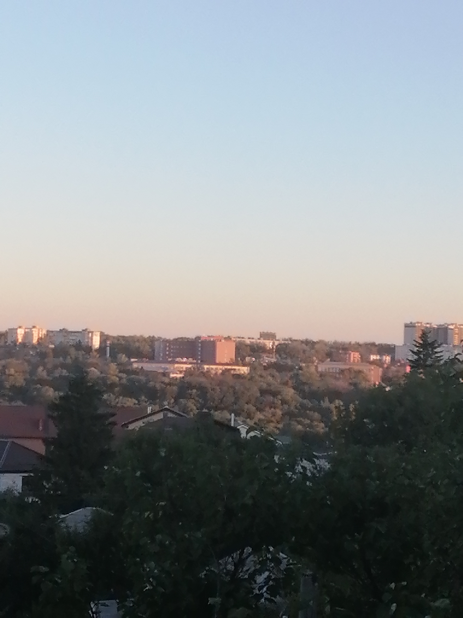 The image captures a view of the city of Rostov-on-Don, Russia, during what appears to be either sunrise or sunset, given the soft, warm light and the clear sky. In the foreground, the tops of trees and some rooftops of houses are visible. The middle ground is dominated by a dense canopy of trees interspersed with various buildings. Among these structures are multi-story apartment buildings and some smaller, more commercial-looking buildings. One notable building in the distance appears to be a large, light-colored structure with a tower. The background reveals more of the city's skyline, characterized by more apartment blocks and a few taller structures, including what looks like a modern residential building with a distinctive facade. The overall impression is of a suburban or semi-urban landscape with a mix of residential areas and larger city buildings, nestled among natural greenery, under a calm, lightly colored sky. There are no people or specific actions depicted in the image.