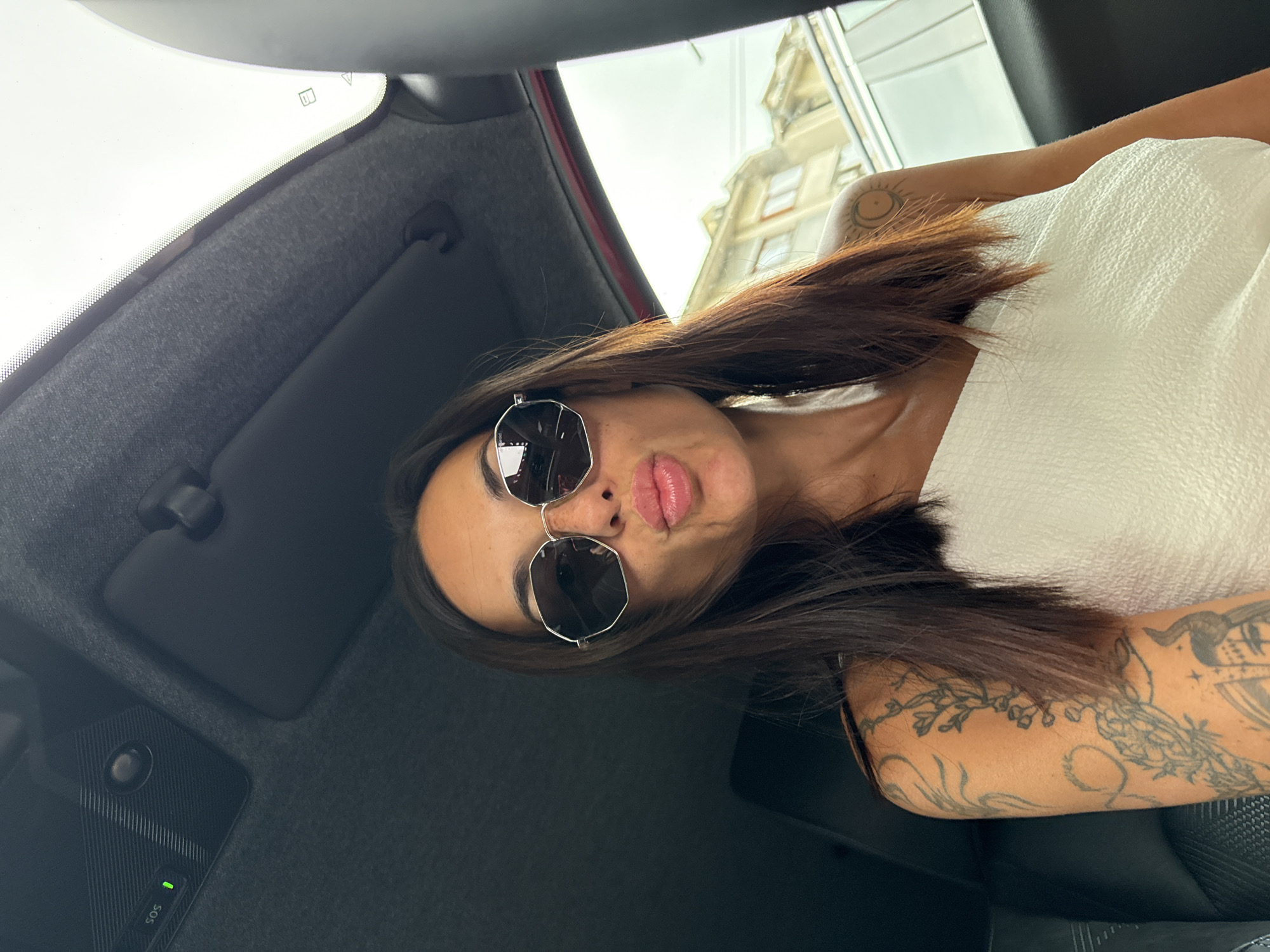 A woman is taking a selfie inside a car. She is wearing sunglasses and making a duck face. She has tattoos on her arms. The car's interior is visible, including the roof lining and a portion of the dashboard with an SOS button. Outside the car, a building and a street can be seen through the window. The scene is likely in the city of Pleven, Bulgaria, as indicated by the architecture and the presence of a car that appears to be a model common in Europe.