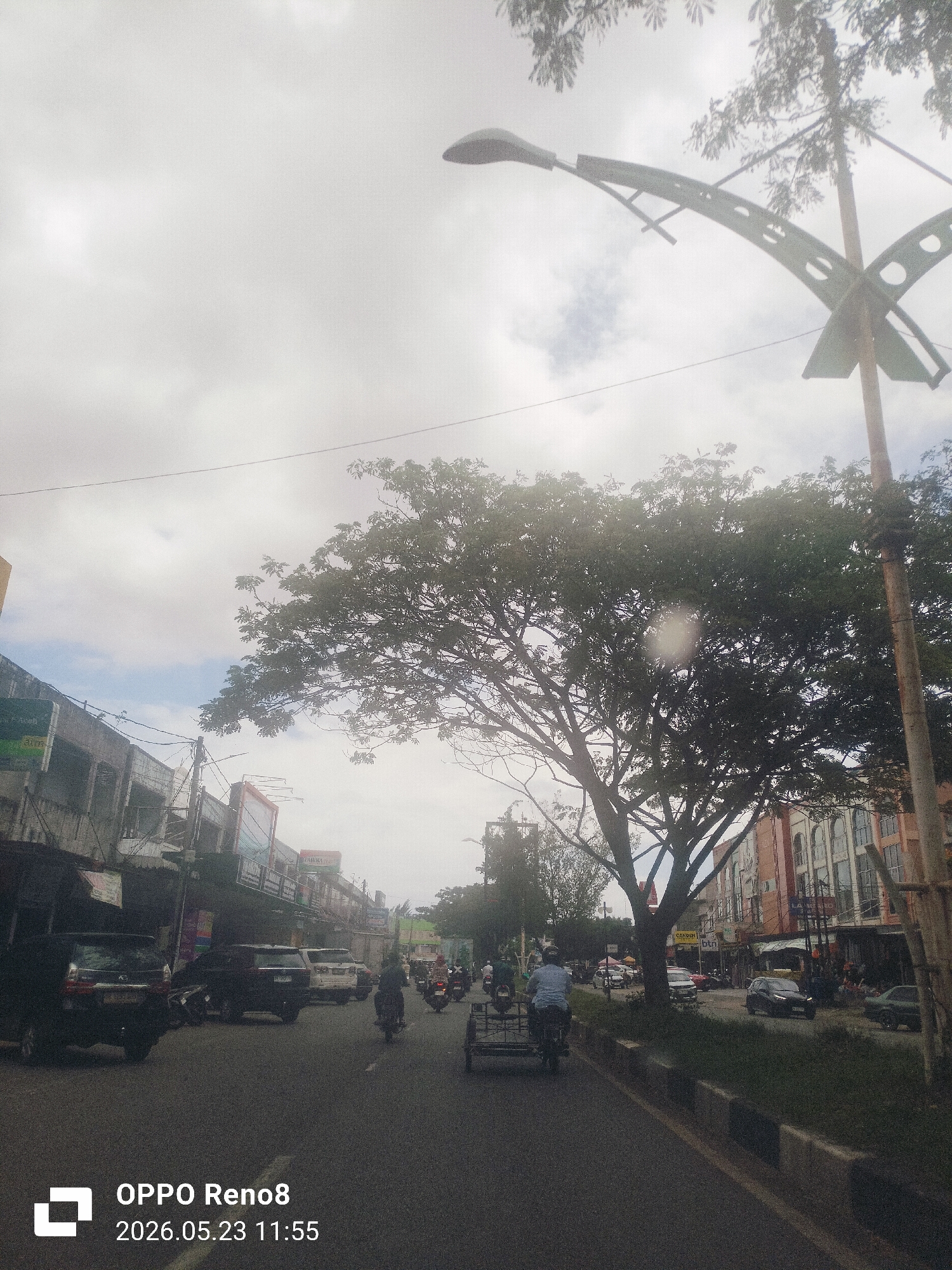 The image depicts a street scene in Lambaro, Indonesia. The photo was taken on May 23, 2026, at 11:55 AM. The sky is overcast. A wide road is in the foreground. On the left side of the road are several buildings with shops and signage. Various vehicles, including cars and motorcycles, are moving along the road. A person is riding a motorcycle with a sidecar. A large tree dominates the center of the image, partially obscuring buildings on the right side of the road. A decorative streetlight stands on the right side. Buildings with architectural details are visible on the right side of the road.
