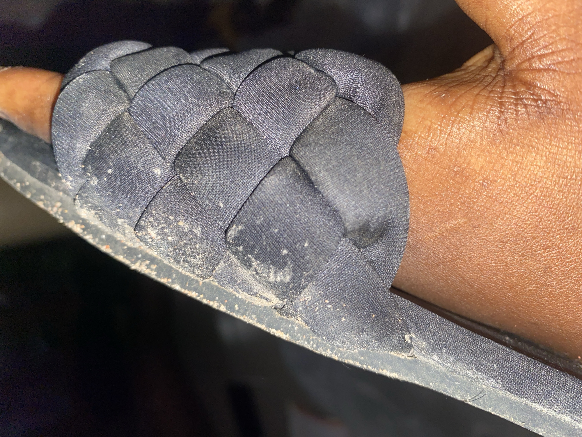 The image shows a close-up view of a person's hand, with the thumb and index finger inserted into a dark blue, padded sandal strap. The strap has a woven or braided design. The sandal appears to be dusty, with visible specks of dirt on the fabric and along the edge of the sole. The hand is brown-skinned and shows some texture and creases. The background is dark and out of focus, making it difficult to discern specific details of the environment. The image was taken in Jauro Yinu, Nigeria.