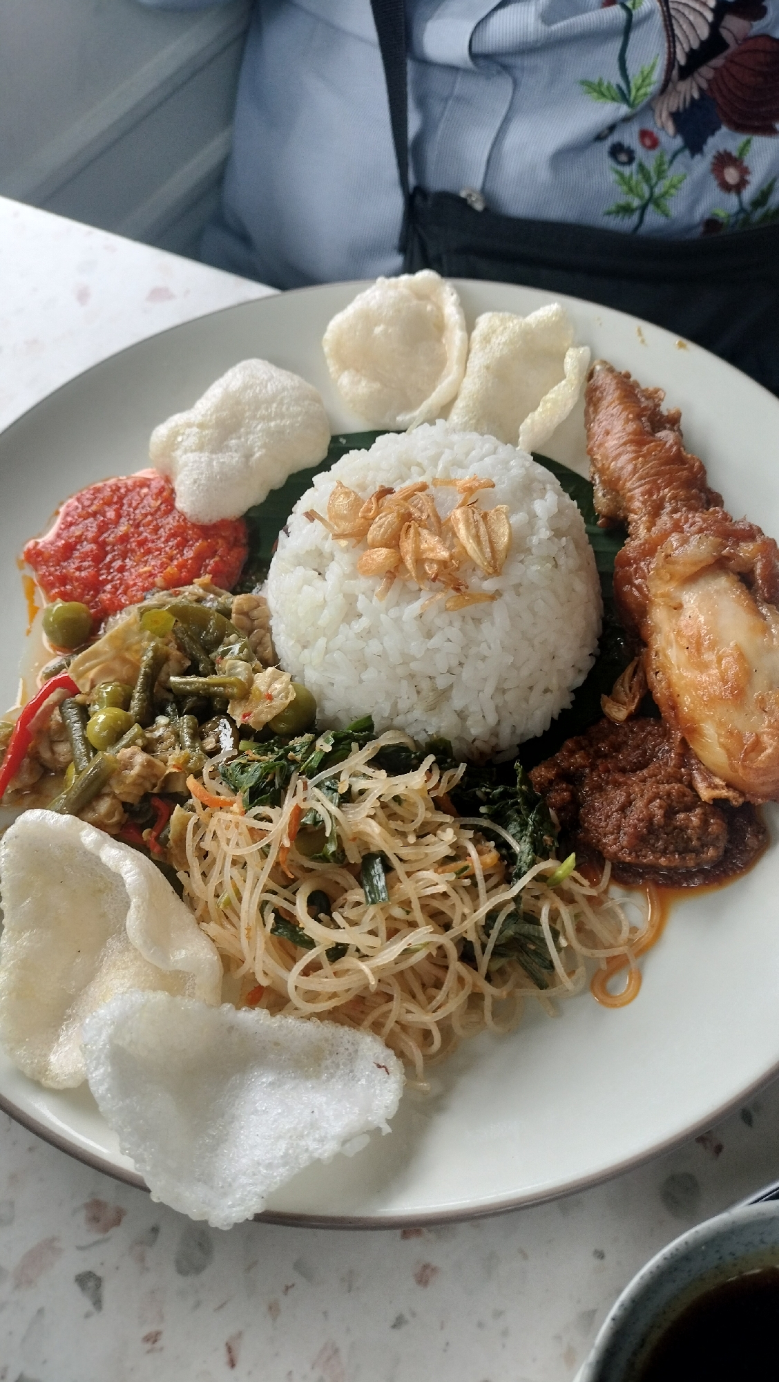 A close-up, high-angle shot shows a plate of food on a light-colored, speckled surface. The plate is white and filled with rice, chicken, vegetables, and crackers. A mound of white rice sits in the center, topped with fried shallots. To the right of the rice is a piece of roasted chicken with crispy skin. Below the rice and chicken are various side dishes: a reddish chili paste, green beans and peas in a sauce, and a pile of thin noodles with mixed vegetables. Several white, puffy crackers are placed around the edge of the plate. A person's torso is visible in the background, wearing a light blue shirt with an embroidered floral pattern. The environment appears to be indoors, possibly a restaurant, with a hint of a window or wall on the left.