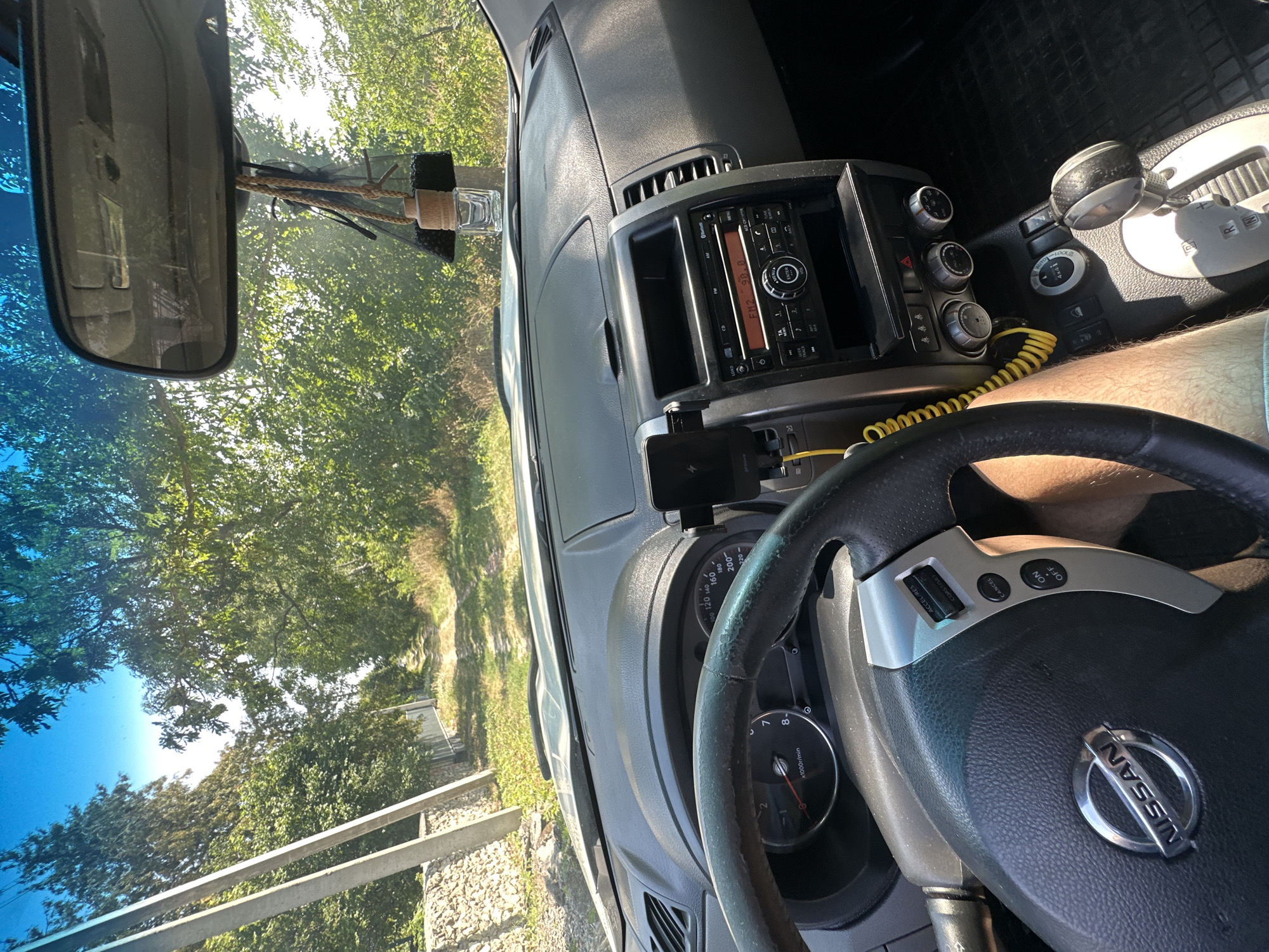The image is a first-person view from inside a Nissan SUV driving on a dirt road in Makiv, Ukraine. The dashboard, steering wheel, and center console are visible. Through the windshield, a dirt road lined with trees and power lines can be seen. The sky is clear and blue, indicating a sunny day. The car's interior appears to be in good condition, with a mobile phone holder attached to the dashboard and an air freshener hanging from the rearview mirror. The driver's leg is visible in the lower right corner of the image, suggesting they are driving. The scene depicts a rural environment, likely outside the main town area of Makiv.