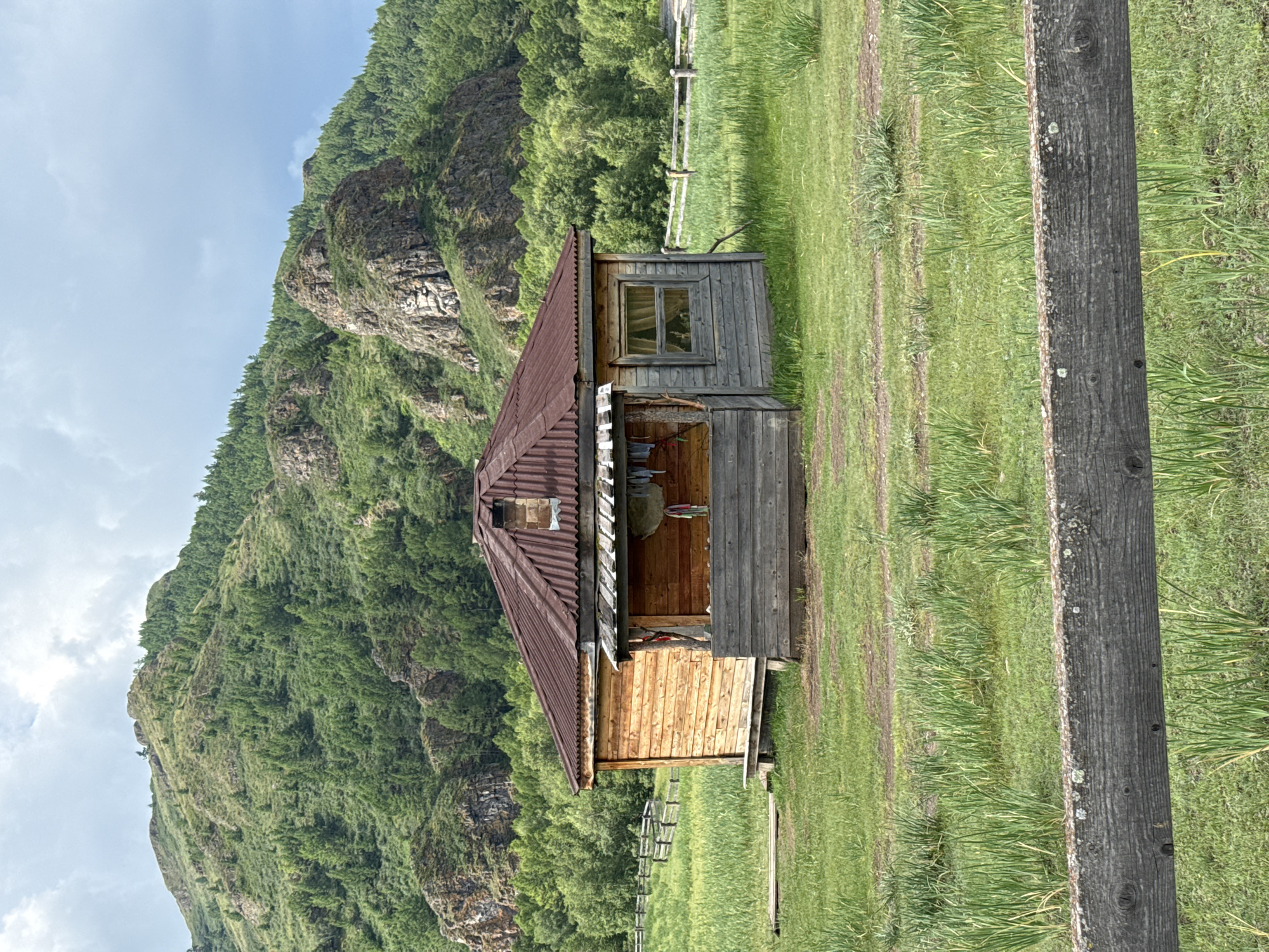 The image depicts a wooden cabin situated in a grassy field in Kazanovka, Russia. The cabin has a corrugated metal roof and is constructed from horizontal wooden planks. A porch area with a wooden railing is attached to the front. Behind the cabin, a lush green mountain covered in trees rises, with rocky outcrops visible. A wooden fence runs horizontally across the field in the middle ground, and another fence is partially visible in the foreground. The sky is partly cloudy. There are no people or animals visible in the scene.