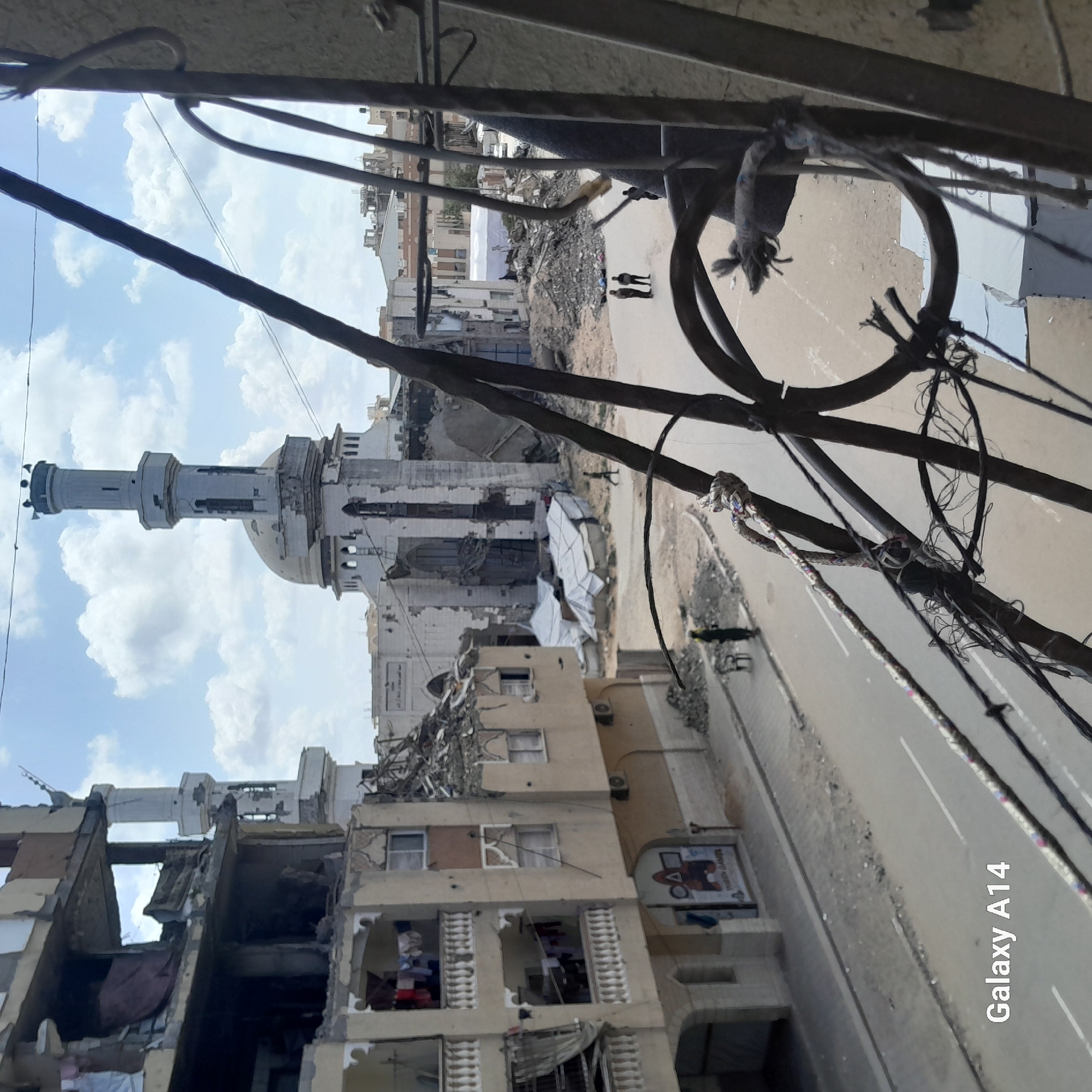 The image shows severe urban devastation, likely in a war zone, in the city of Unknown, Unknown. A large mosque with a minaret is visible in the background, heavily damaged. Buildings in the foreground are also severely damaged, with collapsed structures and debris. Power lines hang haphazardly, and two figures are walking in the street, their backs to the viewer.