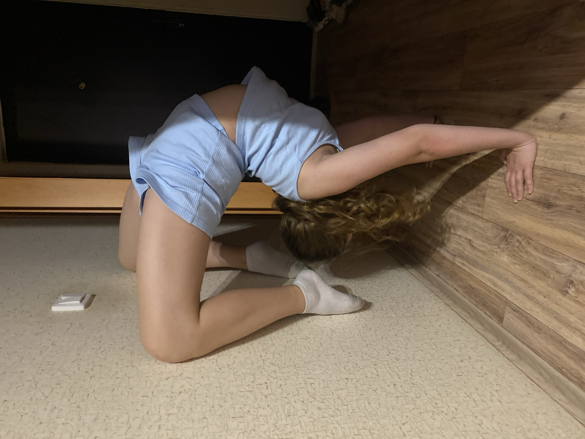 A female individual is positioned in a contorted backbend pose between a wooden wall and a dark, low-hanging structure, possibly a bed frame or other furniture. She is wearing a light blue, short-sleeved shirt and matching shorts, along with white socks. Her arms are extended, with one hand touching the wall. Her face is obscured by her curly, blonde hair. The background shows a wooden floor and wall. A small, white, rectangular object is present on the floor near her knees. The location is indoors.