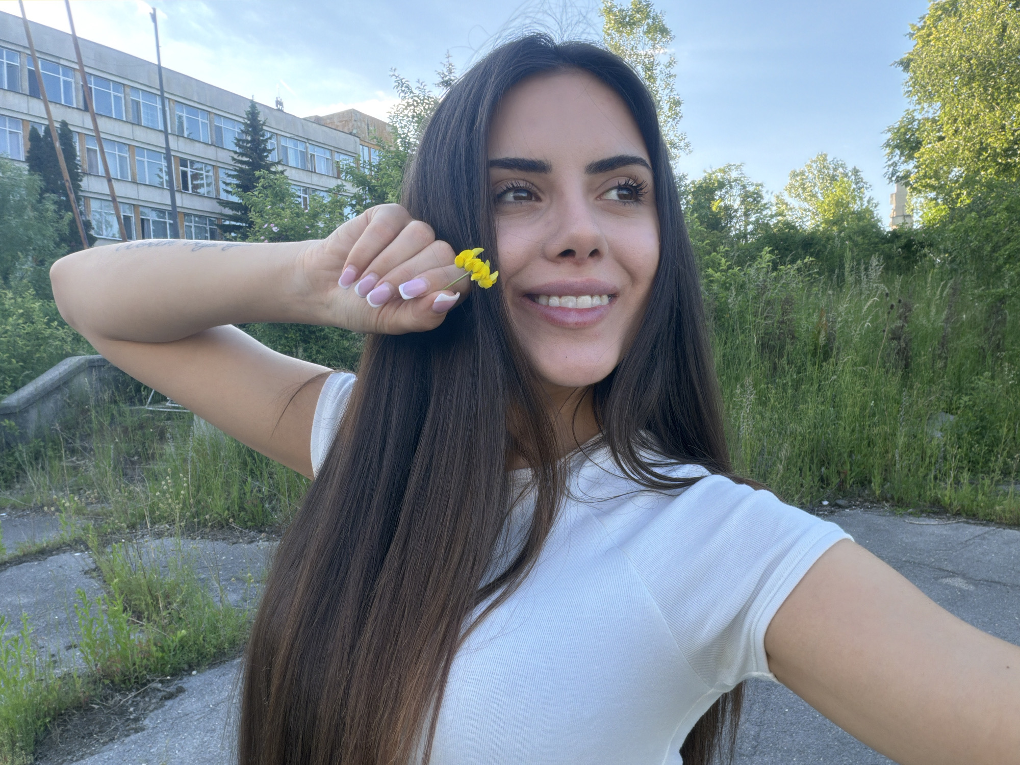 A young adult woman poses for a selfie outdoors. She has long, dark hair and wears a white t-shirt. She is holding a small yellow flower near her face with her right hand. Her nails are painted with a white French manicure. Her right arm is extended, presumably holding the camera.
In the background, there is a large, older building with many windows, suggesting a school or office building. The surroundings appear overgrown with grass and weeds, with trees and bushes filling the scene. The cracked and weathered ground suggests a somewhat neglected or abandoned area.
Based on the architecture and general setting, the location could be in Sofia, Bulgaria. More specifically, it looks like a place on the outskirts of the city near some older institutional buildings or perhaps a university campus. The buildings are characteristic of Soviet-era architecture found in Bulgaria.