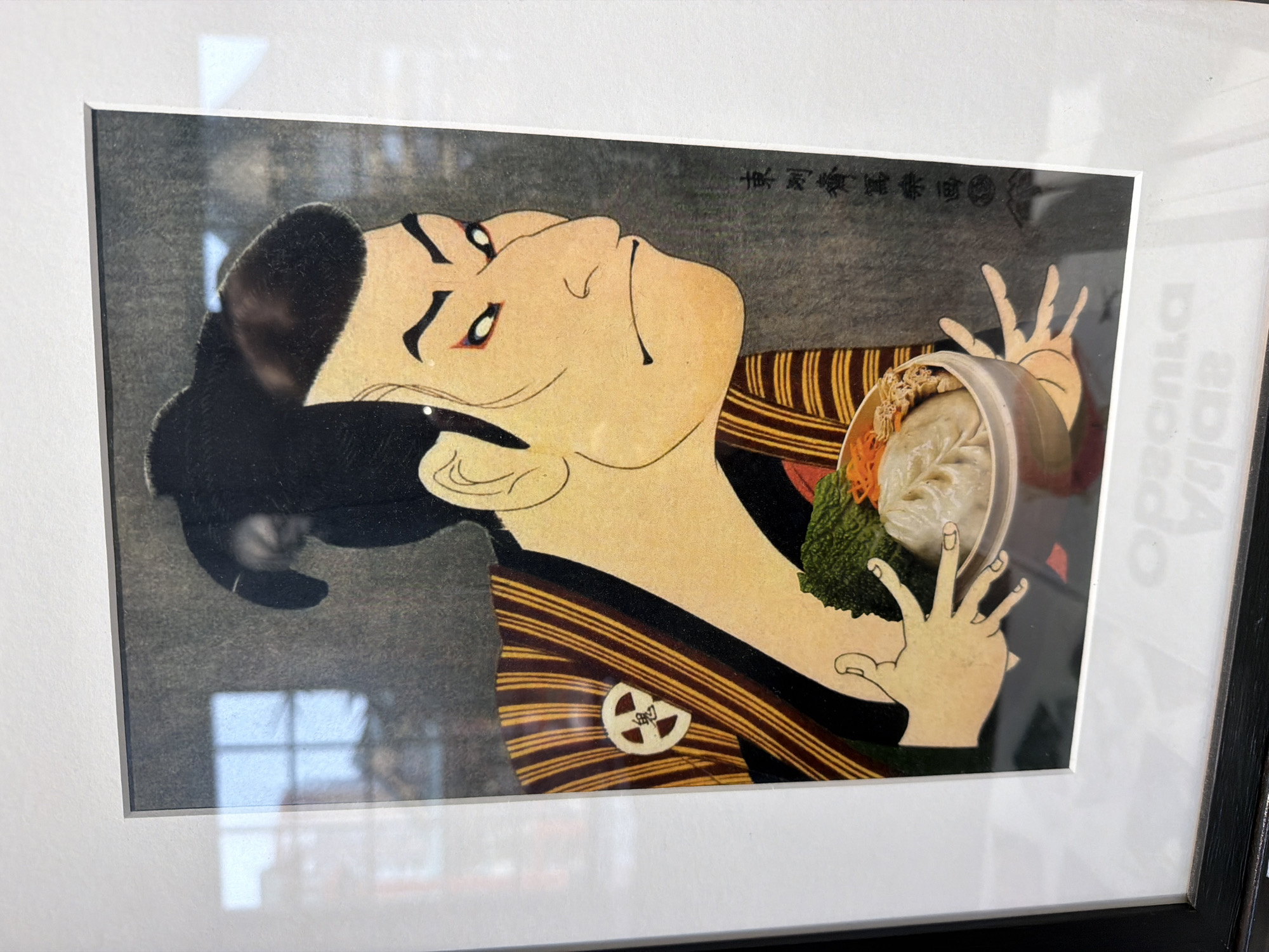 The media file is a photograph of a framed artwork, taken indoors in Khimki, Russia. The artwork itself is a compelling blend of traditional Japanese ukiyo-e style and modern elements.
The central subject of the print is a stylized figure, likely a kabuki actor or a character from classical Japanese theater, depicted with pale skin, dramatically rendered eyes accentuated with red and black makeup, and black hair neatly pulled back into a traditional bun. The figure's expression is intense and theatrical, characteristic of ukiyo-e prints, suggesting a moment of dramatic presentation or profound emotion. This person is adorned in a richly striped garment featuring horizontal yellow and dark brown bands, and a circular white emblem with a dark, stylized symbol is visible on the fabric near the shoulder.
In a striking contemporary twist, the figure is shown holding a clear, modern plastic takeout container filled with food. Inside the container, a large, white, steamed dumpling (resembling a bao or momo) is prominently displayed, garnished with vibrant shredded orange carrots and a dark green leafy vegetable, possibly lettuce or kale. The background of the print is a textured dark grey.
The photograph captures the artwork slightly rotated, giving the impression that the figure within the print is looking upwards. The print is housed in a simple black frame with a wide white matting. Significant reflections on the frame's glass obscure parts of the image, particularly in the corners, with a distinct grid pattern of a window visible in the lower-left reflection.
Visible text includes Japanese characters in the upper right corner of the print, likely a signature or title, and a single Japanese character within the emblem on the figure's clothing. On the right side of the frame, faint, partially reversed text, possibly "Opapoa dja" or similar, is discernible from reflections.