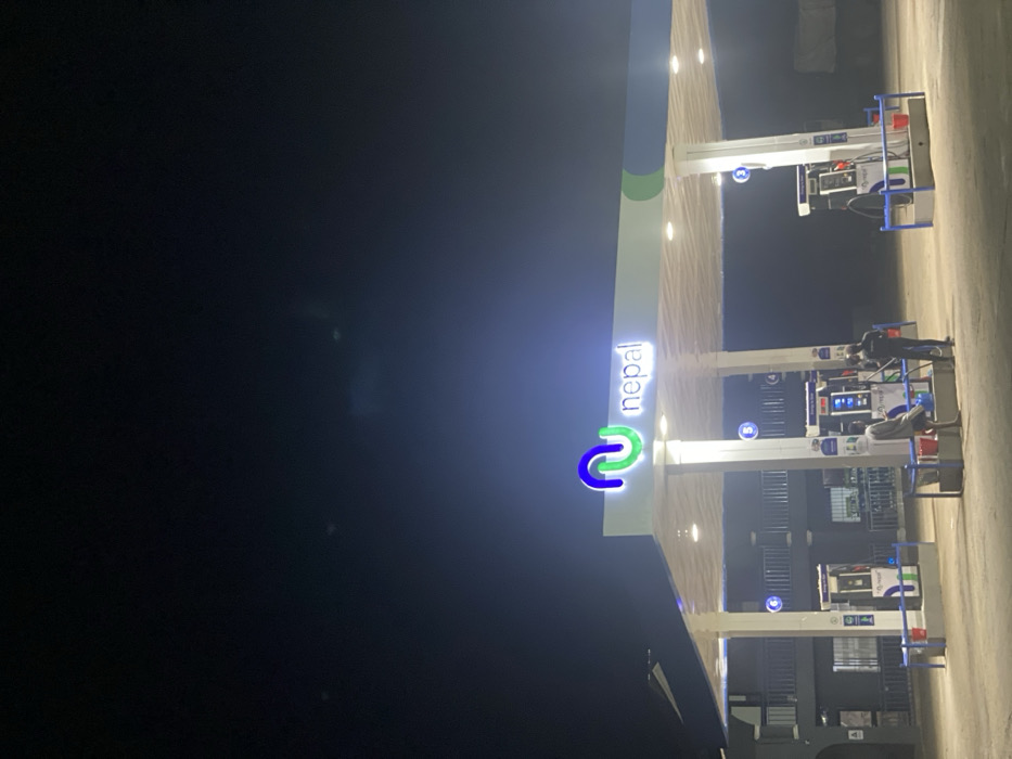 The image displays a nighttime view of a Nepal brand gas station located in Issele Uku, Nigeria. A brightly illuminated canopy, supported by multiple white pillars, features a prominent blue and green "Nepal" logo with white text. Recessed lights are active under the canopy, illuminating the service area. Two fuel dispenser islands are visible. At the leftmost island, two adult males are present. One individual, wearing a light-colored top and dark trousers, is standing near a fuel dispenser with several red jerry cans positioned on the paved ground. The second individual, dressed in dark clothing, is standing adjacent to the first, also in proximity to the fuel dispenser and containers. The actions indicate fuel is being dispensed into the portable containers. To the left of the gas station structure, a multi-story building with balconies and railings is visible, also under illumination. The surrounding environment is dark, consistent with nighttime conditions.