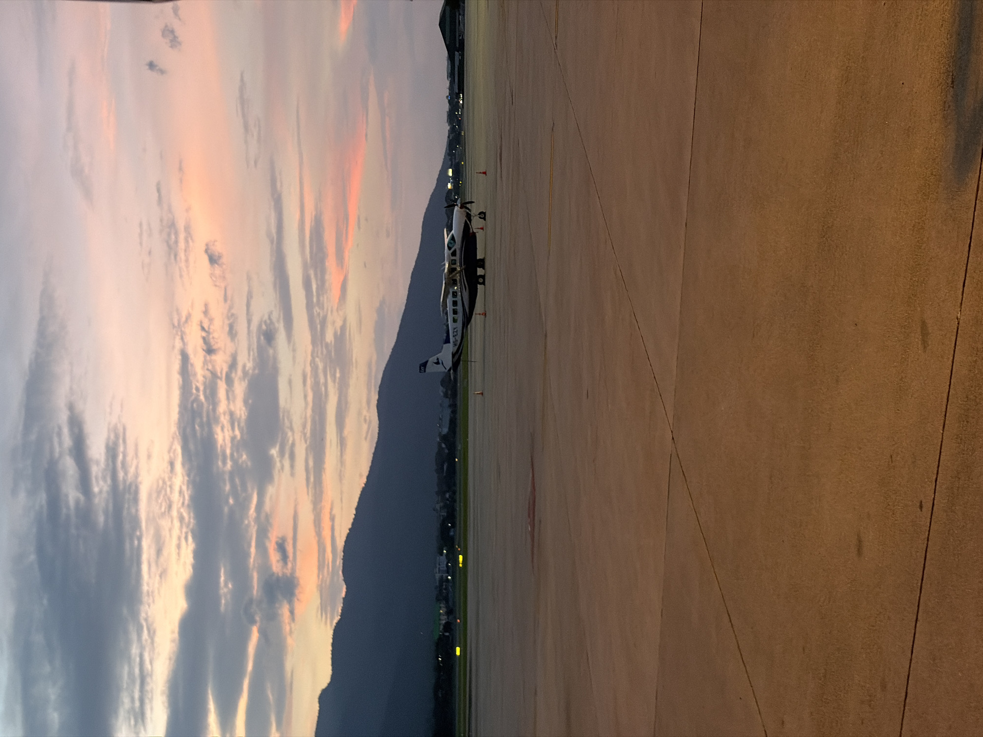 The image captures an airport tarmac at dusk, with a small propeller plane parked in the foreground. The sky is painted with hues of pink, orange, and gray clouds, suggesting either sunrise or sunset. In the background, dark mountains loom, and distant city lights twinkle, hinting at the location of Suthep Town Municipality, Thailand. The tarmac itself is a flat, concrete expanse with visible expansion joints. The plane is white and dark with visible registration markings "HS-EZX" on its fuselage. Several orange traffic cones are placed around the aircraft. The overall scene is serene and still, conveying a sense of quiet anticipation before the night or the start of a new day. There are no people visible in the image.