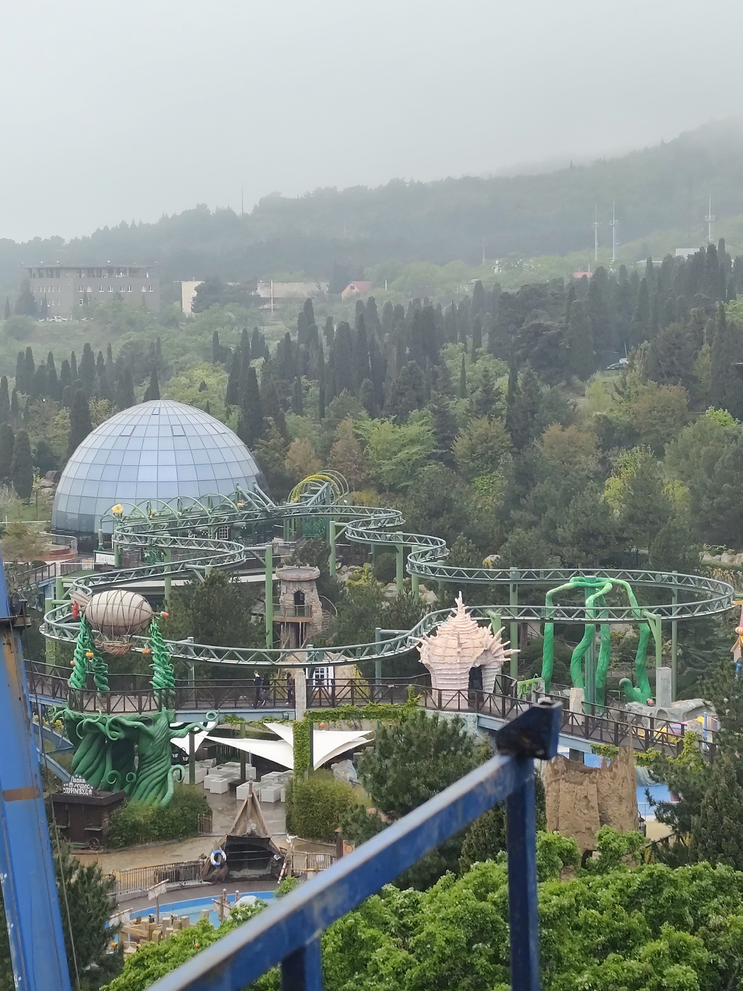 The image depicts a theme park with a roller coaster winding through trees and fantastical structures. Dominating the scene is a large, glass dome structure. The roller coaster's track is green, with themed elements such as giant, green beanstalks and a white, shell-shaped building. People can be seen walking on a bridge connecting the structures. In the background, a building is visible amidst dense trees, and a hazy mountain range fills the horizon. The foreground shows part of a blue structure, likely a ride. The weather appears overcast. The location is Unknown, Unknown.