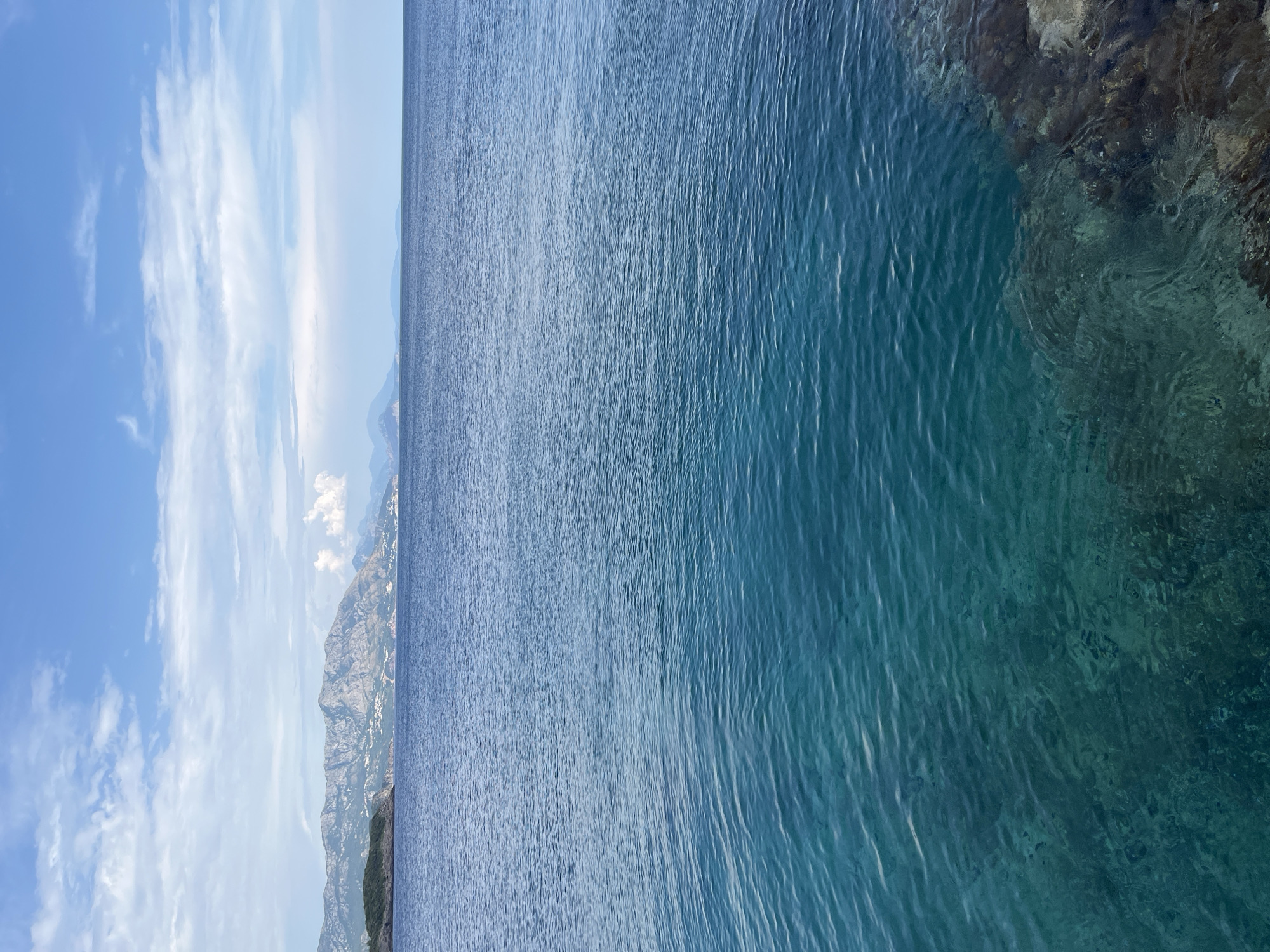The image shows a view of the Adriatic Sea from the coast near Dubraljevina, Montenegro. The water is a clear, deep blue, with lighter turquoise hues visible closer to the shore where it is shallower. The surface of the water has gentle ripples. In the foreground, rocks are visible submerged beneath the clear water, indicating a rocky coastline.
In the distance, a mountainous coastline stretches across the horizon, dotted with buildings. The mountains are covered in vegetation, and their peaks are rugged. The sky above is a bright blue, with scattered white clouds. The overall scene is serene and captures the natural beauty of the Montenegrin coast. There are no people or animals visible in the image.
