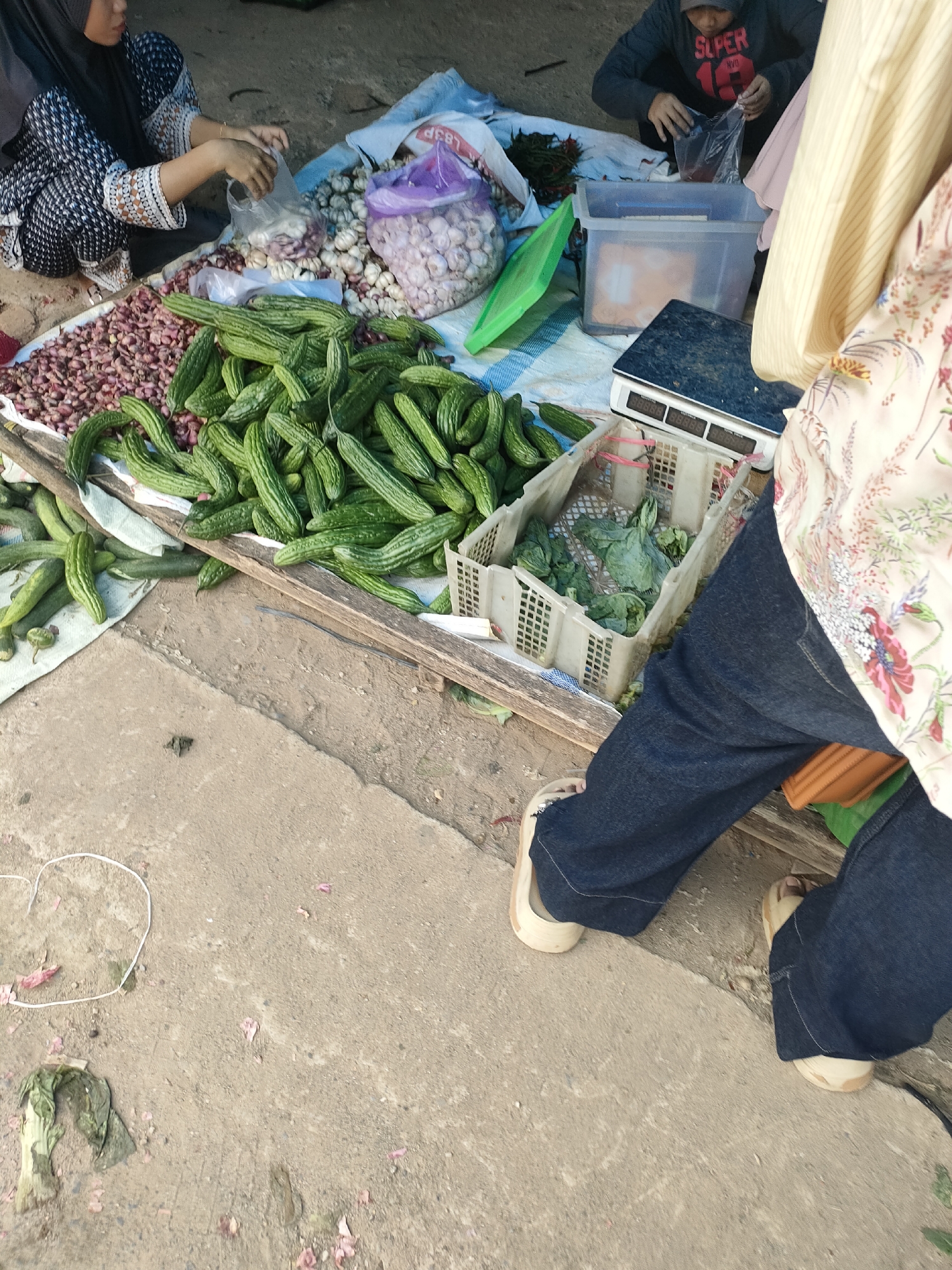 The image captures a market scene in Sukoyoso, Indonesia. In the foreground, a person is wearing dark blue jeans and light-colored sandals. To the left, a woman wearing a black hijab and a patterned dress is seen behind a display of vegetables. Piles of red onions and green bitter gourds are prominently featured. In the background, another person is visible wearing a grey hoodie with the word "SUPER" printed on it, engaged in an activity that involves a plastic bag. Various market items, including garlic and leafy green vegetables in plastic crates, are also present. A scale is visible on a blue tarp. The ground appears to be a mixture of concrete and dirt, with scattered debris.