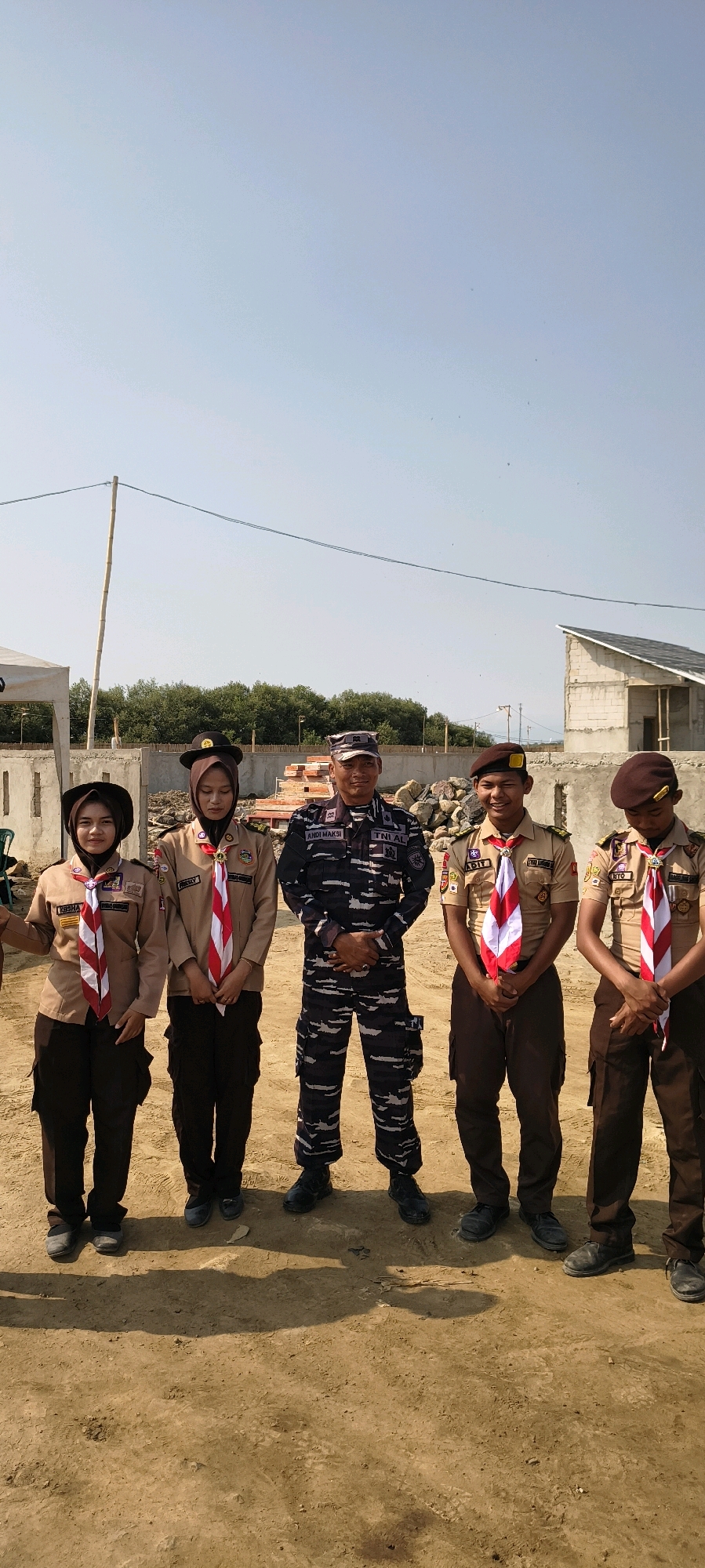 In Gebang, Indonesia, five individuals stand in a line outdoors under a clear sky. On the left, two young women wear scout uniforms in a tan color with brown pants and red and white neckerchiefs. Both have dark hair and are looking towards the camera. The first woman has a black headscarf, and the second wears a brown beret. In the center, a man in a dark blue and black camouflage uniform, identified as "ANDI MAKSI" and "TNI AL," stands with his hands clasped in front of him. To his right, two young men also wear tan scout uniforms with brown pants and red and white neckerchiefs. The first young man has a brown beret and is looking towards the camera with a smile, identified as "ADIT." The second young man, to his right, wears a brown beret and looks downwards. Behind the group, construction materials, including bricks and rocks, are visible, along with a concrete wall and the partially constructed frame of a building with a grey roof. A bamboo pole with wires stretches across the upper background, and trees are seen in the distance. The ground is covered in loose, sandy soil.
