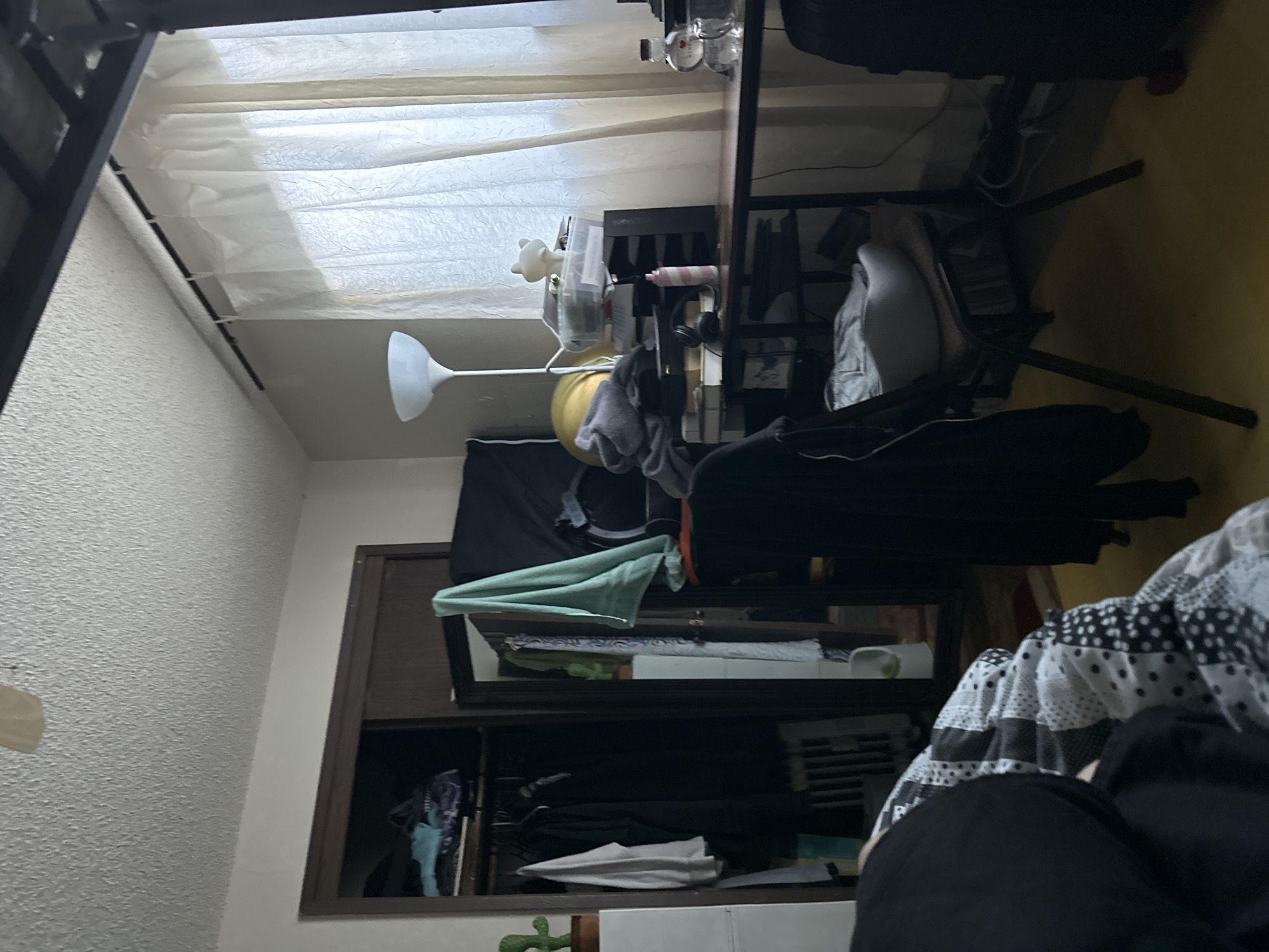 The image depicts a cluttered bedroom. On the left, a closet is partially open, revealing clothes on hangers and shelves with items. A small green cactus figurine sits on the top shelf. To the right of the closet, a desk is positioned against a wall, covered with various objects including a lamp, a plastic bin filled with items, and a pink bottle. A chair with a white cushion is placed in front of the desk. A large window covered by sheer white curtains occupies the background. The floor is a yellow linoleum. In the foreground, a person is lying in bed, covered by a black and white patterned comforter. The overall impression is of a lived-in and somewhat disorganized personal space. The location is not identifiable as Vancouver, United States of America.