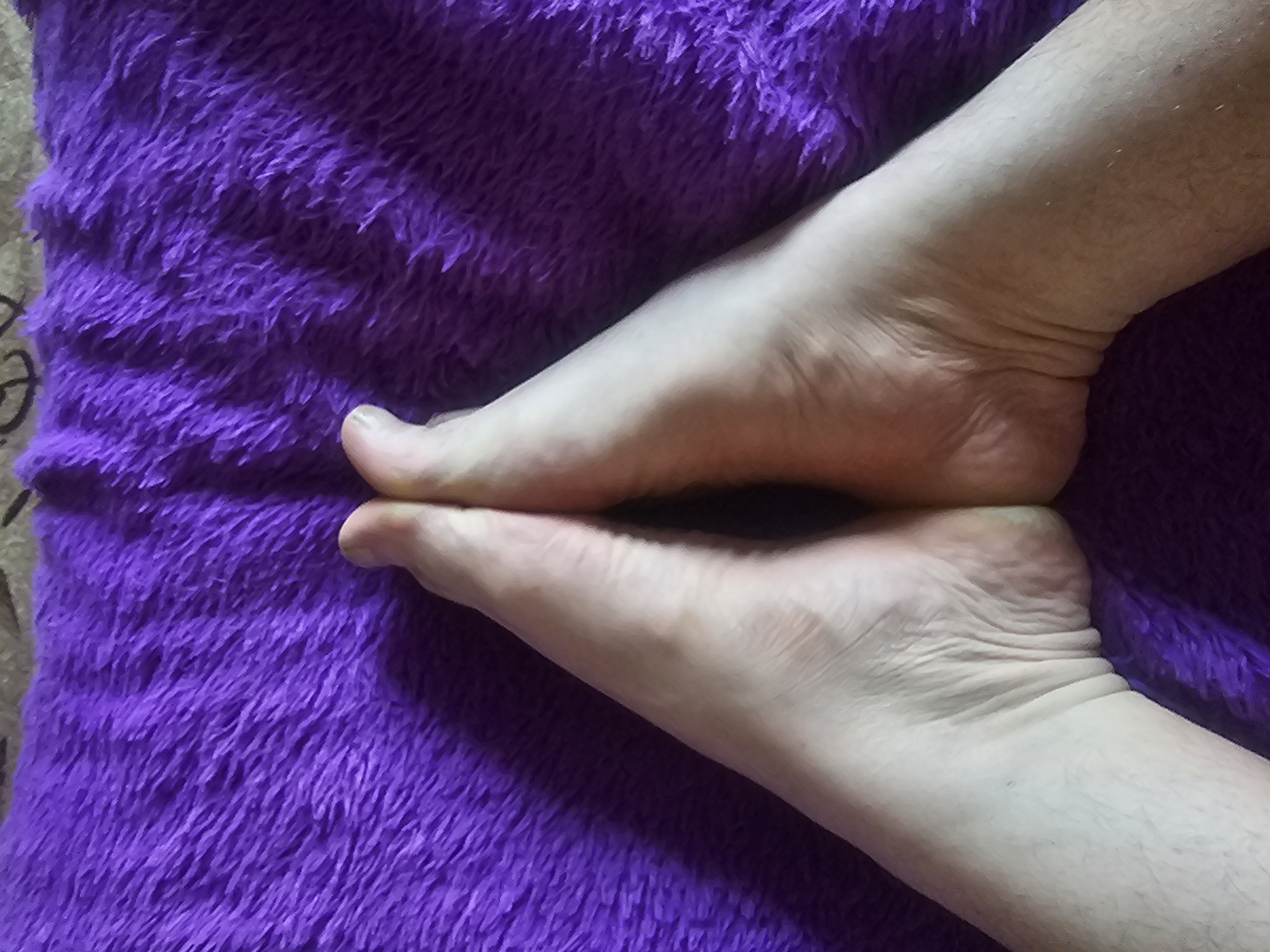 Two bare feet are positioned close together, soles facing upwards, against a textured, vibrant purple surface. The feet are pale, with visible skin creases and some light hair on the ankles and lower legs. The purple surface appears soft and possibly furry, with a dense, short pile. The image is taken from a high angle, looking down at the feet. The location within the city of Таёжное, Russia is not discernible from the visual information provided.