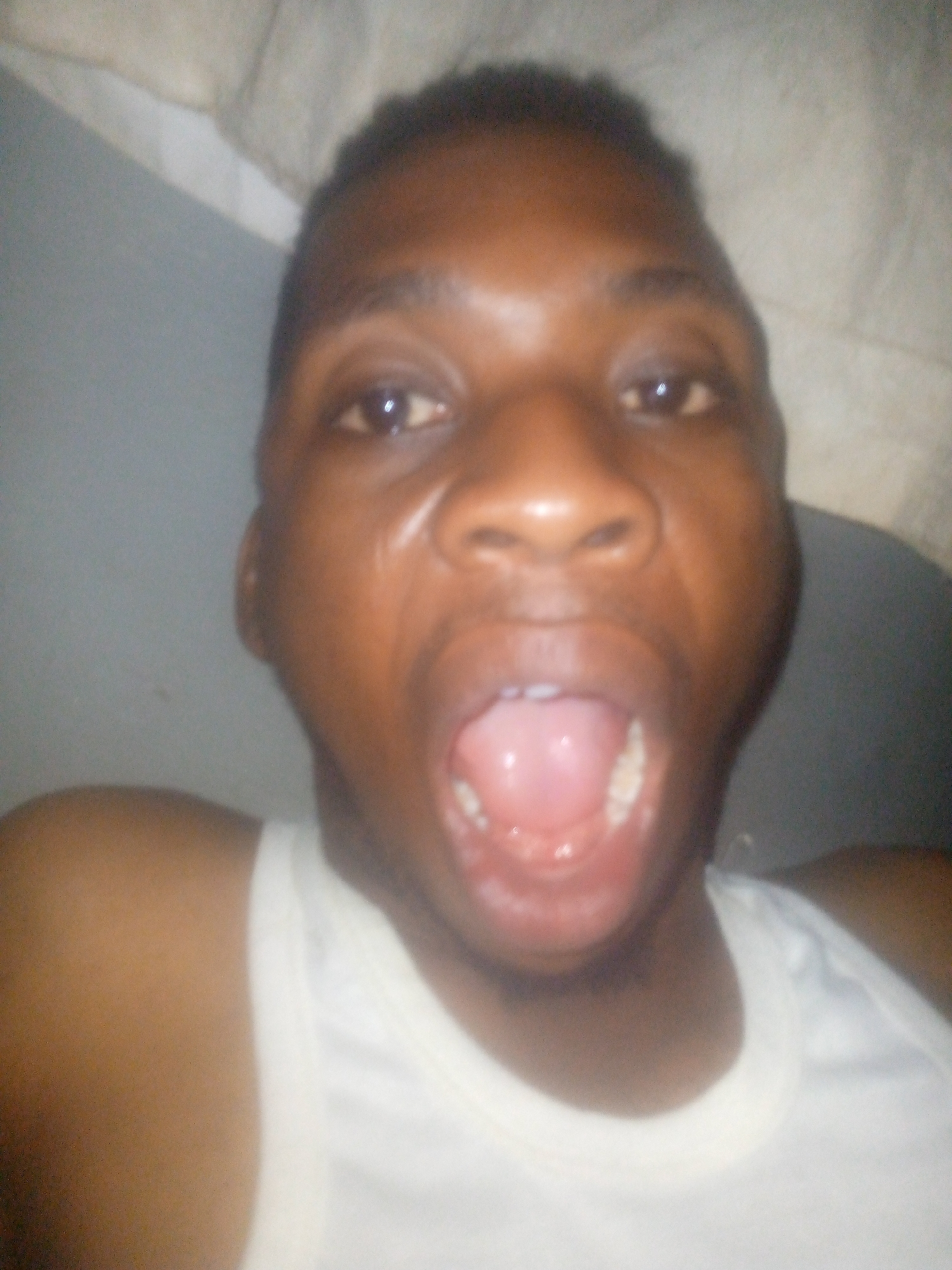 A close-up selfie of a dark-skinned male with an open mouth, revealing teeth and tongue. He is wearing a white tank top. The background consists of a grey wall and a white surface, possibly a sheet or blanket. Location is Unknown, Unknown.
