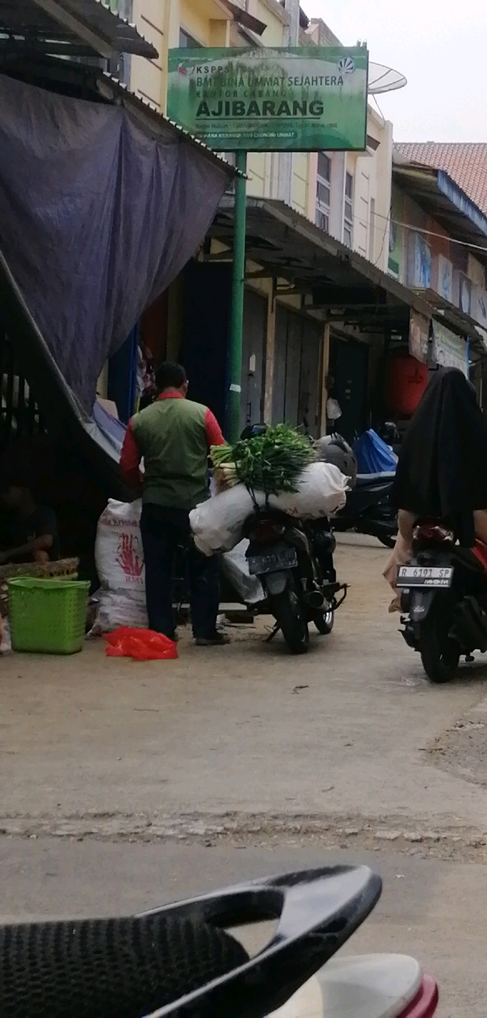The image depicts a street scene in Ajibarang, Indonesia, likely near a market or a small business. A man wearing a red shirt and a green vest is standing in front of a motorcycle loaded with sacks and greenery, possibly vegetables. To his left, a green plastic basket and a red plastic bag are on the ground. A sign for "KSPSPS BM BINA UMMAT SEJAHTERA KANTOR CABANG AJIBARANG" is visible in the background, indicating the location. Another motorcycle, ridden by a person wearing a black hijab, is partially visible on the right side of the frame. The environment appears to be an outdoor, somewhat unkempt urban setting with buildings and closed shop fronts in the background.