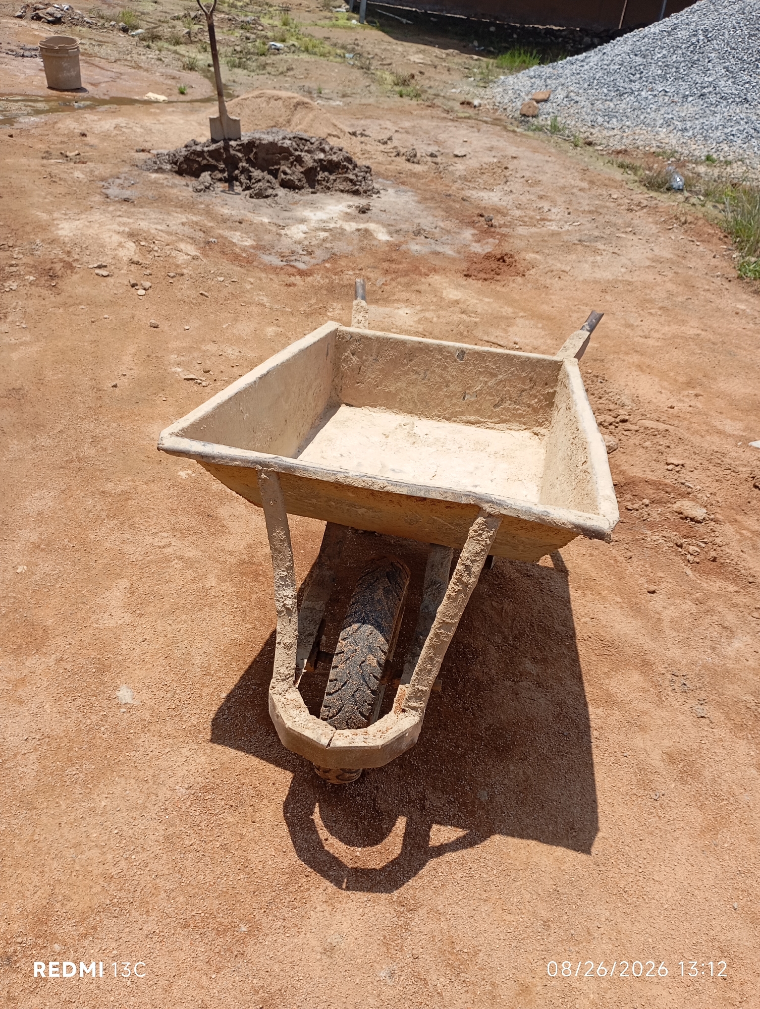The image depicts a construction site in Shendam, Nigeria, characterized by reddish-brown soil. In the foreground, a single, well-worn wheelbarrow made of concrete or a similar material sits on the ground, its basin filled with a light-colored powder, possibly cement or fine sand. The wheelbarrow's frame and tire are visibly coated in dirt and possibly dried mud, indicating recent use. The tire is a dark color with a tread pattern.
In the background, a pile of dark, wet soil is mixed with some sand, and a shovel is partially embedded in it, suggesting ongoing work. To the left, a light-colored bucket or container stands on the damp ground. Further back, a large mound of grey gravel is visible, a common material for construction. The environment appears dry and dusty, typical of a construction area in Nigeria. The lighting suggests it is daytime, with a clear shadow cast by the wheelbarrow. A timestamp in the lower right corner reads "08/26/2026 13:12", and "REDMI 13C" is visible in the lower left corner, indicating the device used for capture. There are no people visible in the frame.