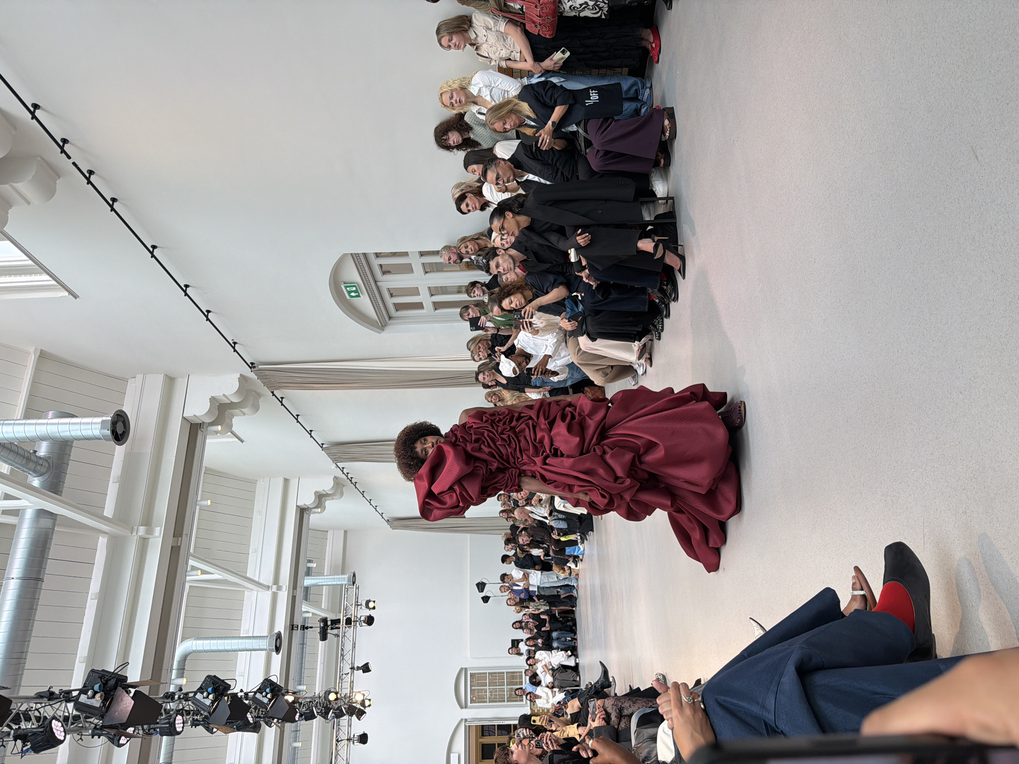 This image captures a fashion show event, possibly held in a modern exhibition space or art gallery, with exposed ceiling structures, industrial ductwork, and track lighting visible. The audience is seated along the sides of a runway, with a model in a voluminous, dark red couture dress walking down the center. The model has an afro hairstyle and is the central focus of the image. The spectators are a diverse group, many of whom are holding up smartphones to take pictures or videos of the show. The overall atmosphere appears to be one of a professional fashion event. The architectural details and lighting suggest a contemporary venue. The city of Utrecht, Netherlands is not explicitly depicted in the image, and there are no landmarks or context that would confirm this specific location.