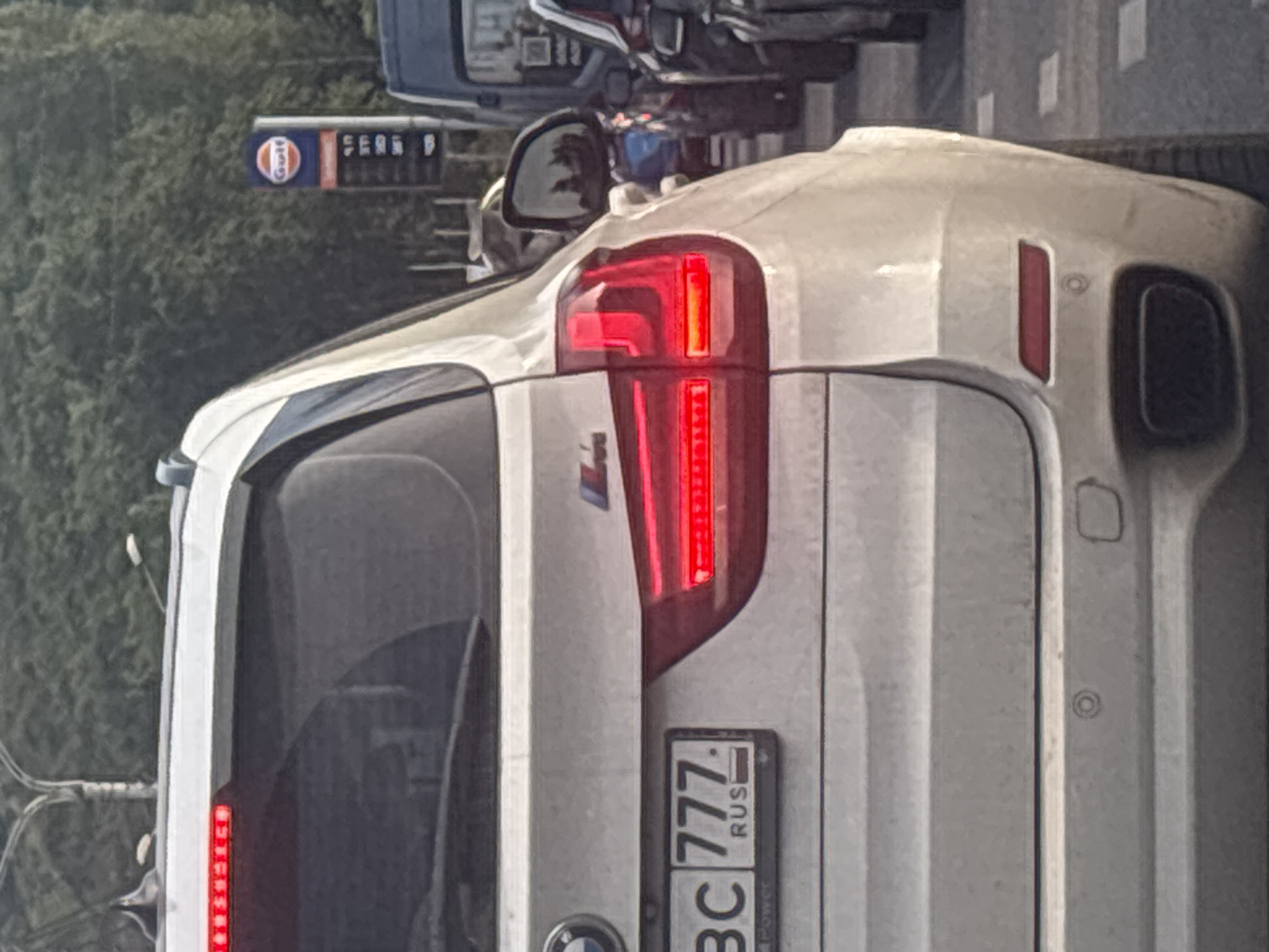 The image shows the rear of a white car, possibly a BMW, parked on a street. The car's taillights are illuminated, suggesting it is either dusk or nighttime, or that the brake lights are on. The car is positioned vertically in the frame. In the background, a Gulf gas station sign is visible, with the Gulf logo and what appears to be fuel prices displayed. There is also a reflection in the car's bumper, hinting at other vehicles or structures nearby. A license plate on the car reads "BC 777 RUS". The overall scene appears to be urban, with hints of greenery in the background, possibly trees. The weather is not discernible.