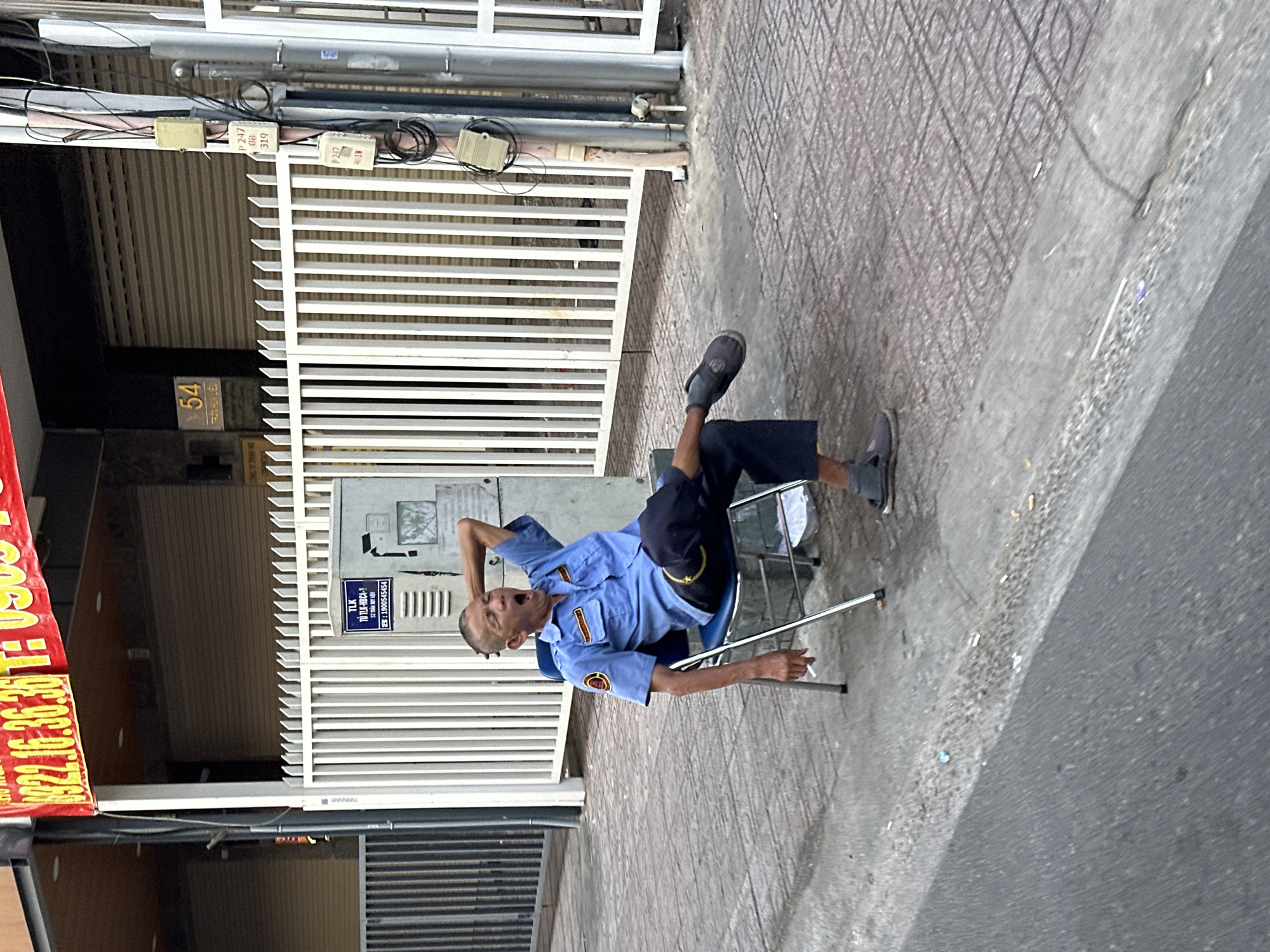 A security guard is sitting on a chair in the city of Thủ Đức, Vietnam. He is wearing a blue uniform and appears to be yawning, stretching, or shouting. He is holding a cigarette and there are numerous cigarette butts on the pavement next to his chair. Behind him, there is a white gate and a building with signage on it, including the address number 54, as well as a red sign with the phone number 0922.16.36.36. A blue sign on the building states: TLK TÙ TLK-HBC4-1 52 TRẤN HUY LIỆU SER: 1900545454. Above the gate, there are electrical boxes and wires. The street is paved.