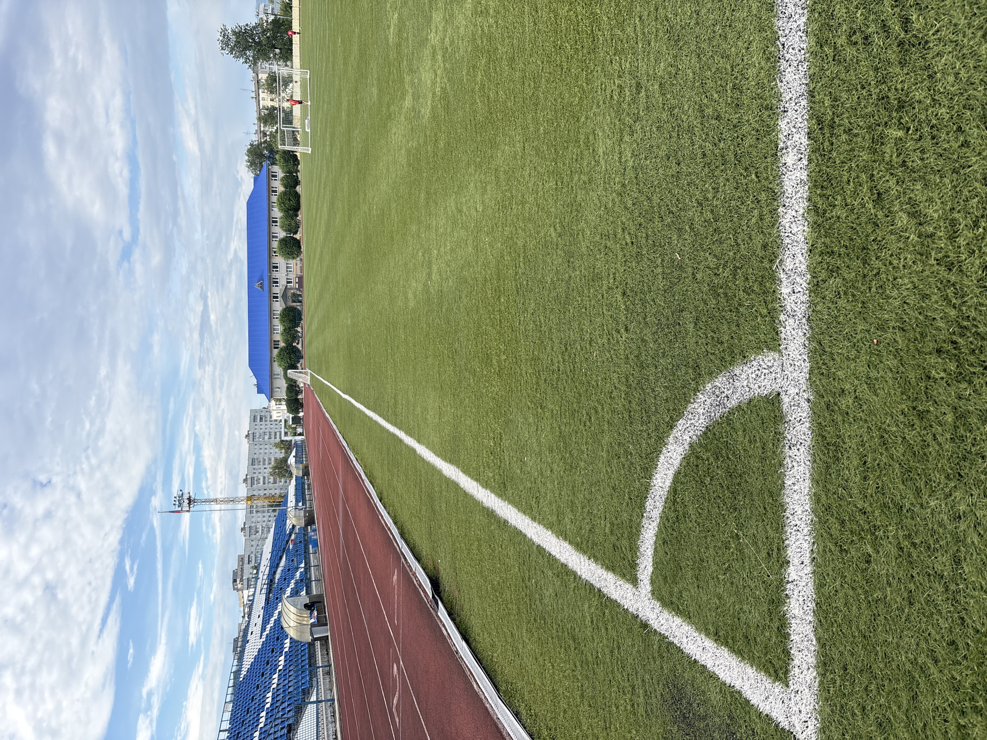 This media file shows a wide-angle view of an outdoor stadium. The foreground features a close-up of a green artificial turf soccer field with white lines marking the boundaries of the field, including a corner arc. To the left of the field is a red running track. Beyond the track are rows of blue stadium seats, some of which are empty. In the background, there is a multi-story building with a bright blue roof, as well as other buildings and trees. A tall lighting tower is also visible. The sky is blue with scattered white clouds. The image appears to be taken during the daytime. The scene is set in Chaikovsky, Russia, as indicated by the location context. There are no discernible people actively engaged in any activity within the frame, though the presence of a soccer field and stadium suggests a sporting event might have taken place or is planned. The weather appears to be fair.