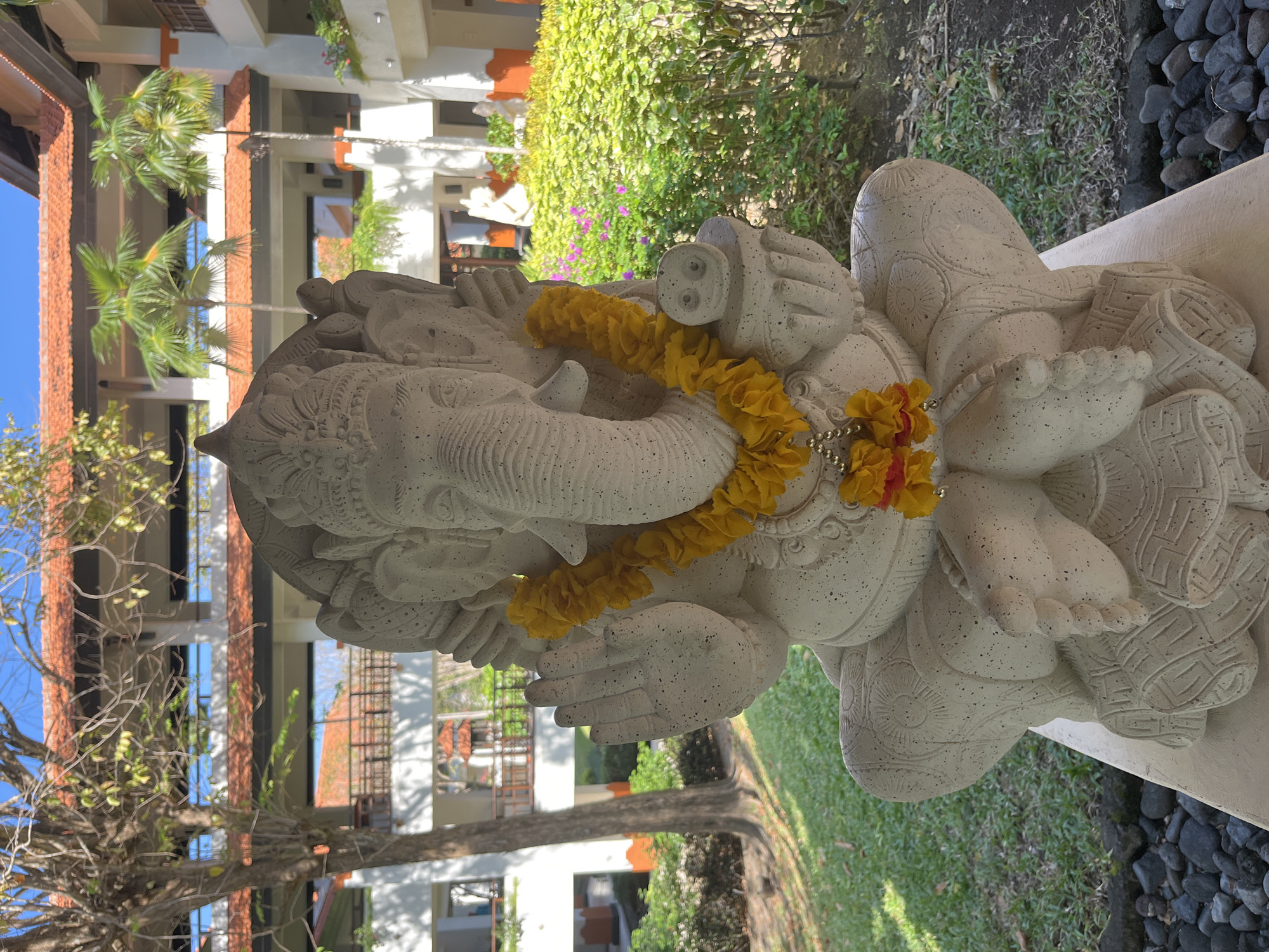 A stone statue of Ganesha, adorned with a yellow floral garland, is prominently displayed in an outdoor courtyard. The statue is seated with its legs crossed and hands raised in a gesture of blessing. Behind the statue, a multi-story building with red-tiled roofs and white facades is visible, featuring balconies and walkways. Lush green foliage and palm trees surround the building, suggesting a tropical or subtropical environment. The ground in front of the statue is covered with grass and a border of dark stones. The sky is clear and blue. The scene appears to be in a resort or a public garden area, likely in Kuta Selatan, Indonesia, known for its numerous hotels and lush landscapes.