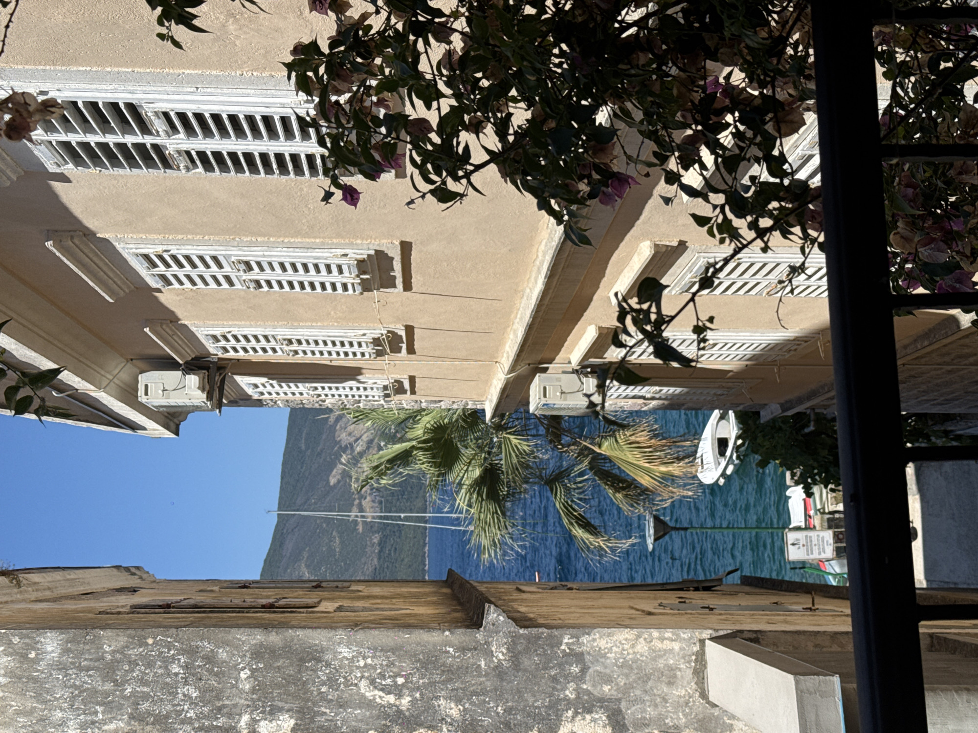 The image depicts a view looking down a narrow street towards the sea. On the left, a weathered stone building with visible window frames stands tall. On the right, a building with a tan facade features white-slatted shutters on its windows and an air conditioning unit mounted outside. The sea is a vibrant blue, with a sailboat mast visible in the distance and a small white boat moored closer to shore. A palm tree with green fronds partially obstructs the view of the water and the sailboat. Lush green foliage with some purple flowers is in the foreground on the right side of the image. The scene is illuminated by bright sunlight, casting shadows.