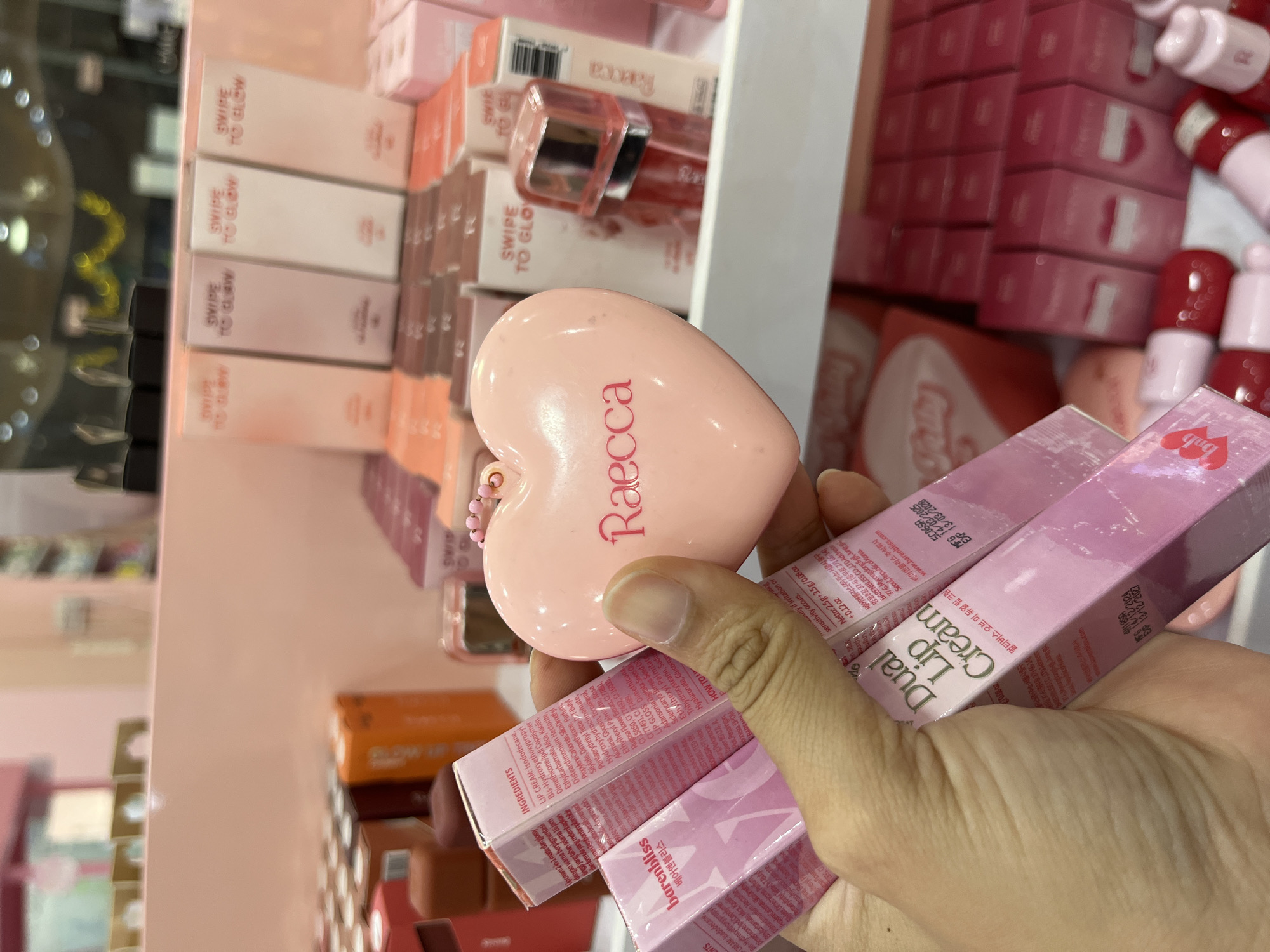 In a retail environment with pink shelving and displays, a person's hand holds several pink lipstick tubes and a pink heart-shaped compact. The lipstick tubes are branded "barenbliss" and "Dual Lip Cream," with Korean text visible on their packaging. The heart-shaped compact has the brand name "Raecca" printed in pink. In the background, rows of pink and orange boxes are displayed on shelves, along with other cosmetic products, including small red lipstick tubes. The overall impression is a cosmetics store aisle, likely in a city like Lhokseumawe, Indonesia, given the context provided.