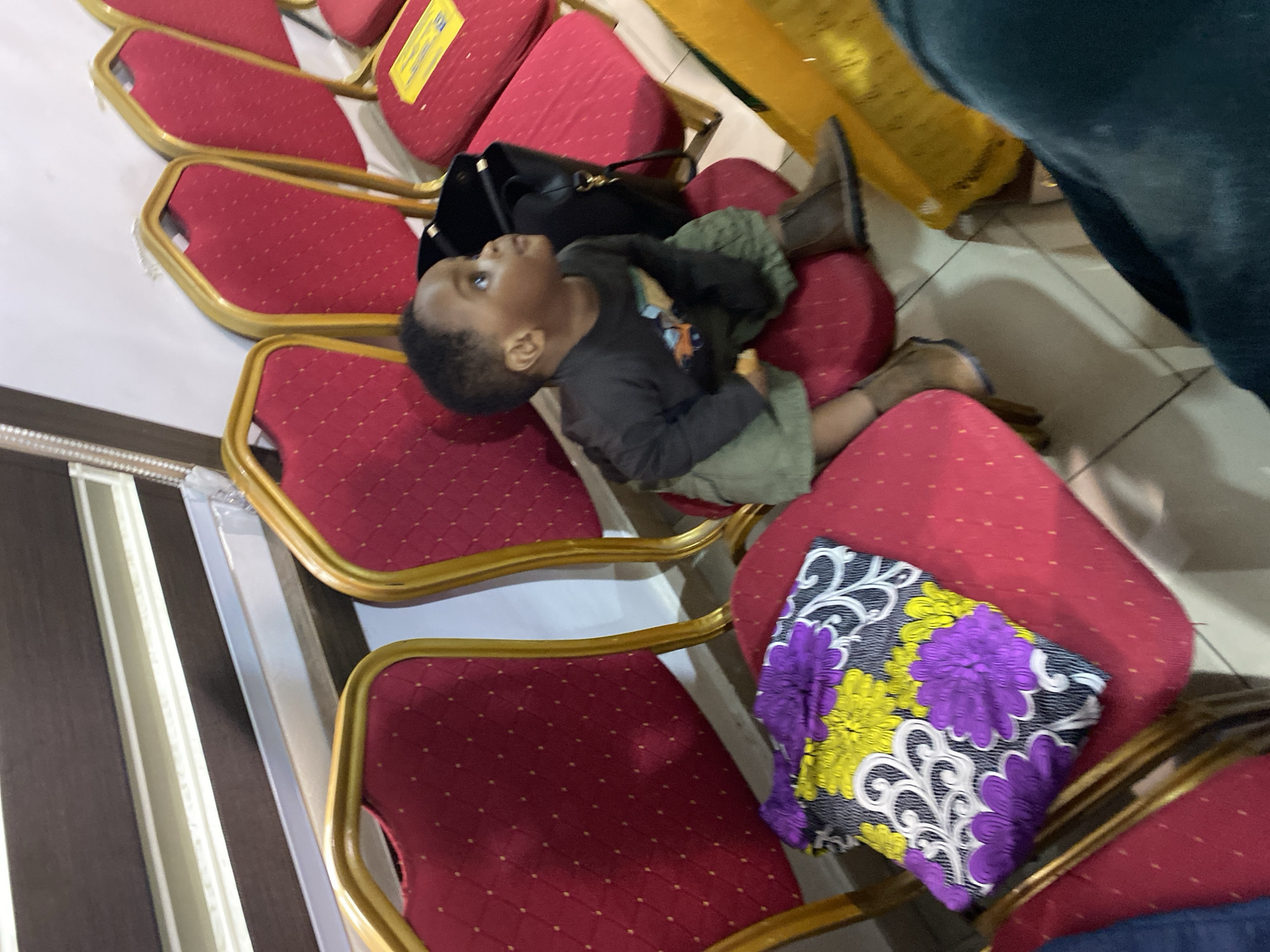 A child rests across several red chairs in an indoor setting. The chairs have gold frames and are arranged in a row, suggesting a waiting area or meeting room. The child is wearing a long-sleeved shirt and olive-green pants, with shoes on. A black handbag is next to the child. There is also a colorful floral pillow on a chair nearby. The setting appears to be inside a public space, possibly in Ado, Nigeria.