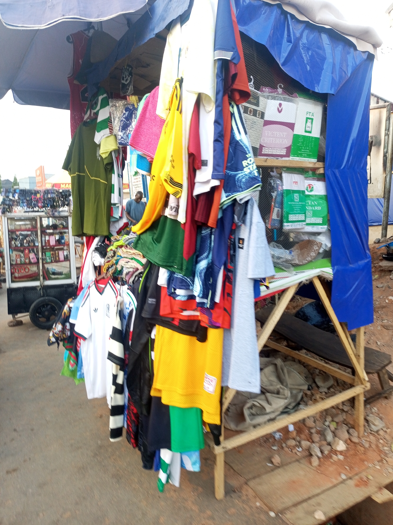 The image depicts an outdoor retail scene in Gombe, Nigeria, likely a market or street vendor setup. Garments, including sports jerseys, t-shirts, and possibly towels, are hanging on display, creating a dense collection of colors and fabrics. The vendor's stall appears makeshift, constructed from wood with a blue tarpaulin roof for shade. Inside the stall, boxes labeled "FTA" are visible. A separate, wheeled cart with glass display cases stands nearby, containing smaller items like cosmetics or accessories. A man is seen standing in the background, possibly the vendor or a customer. The ground is paved, but littered with small rocks and debris near the stall's base.