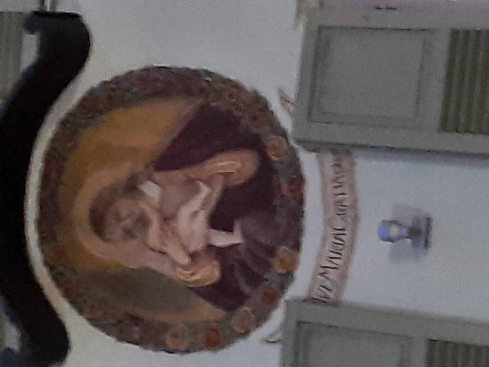 A religious fresco, depicting Mary holding the infant Jesus, is centrally located above a banner inscribed with German text. The fresco is framed by a circular motif of flowers. Below the fresco, a small, stylized bust of a figure is mounted on the wall. The fresco and bust are situated on a white wall, flanked by two light-colored framed panels, suggesting an interior setting possibly within a church or other religious building in Rosenheim, Germany.