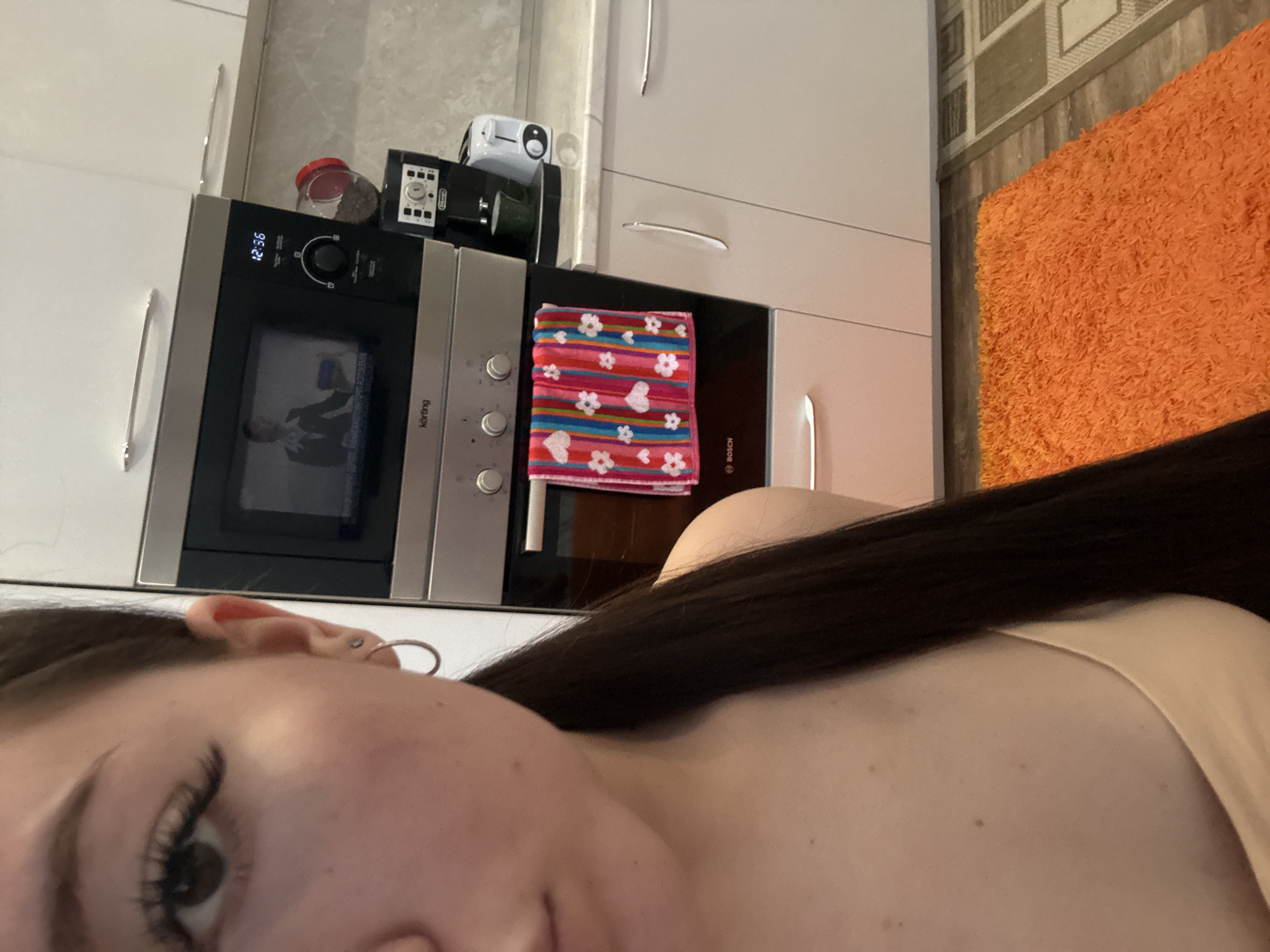 The image is a close-up, rotated selfie showing a person and a kitchen background. The subject is a female with long dark hair, visible on the left side of the frame. Her right eye with prominent eyelashes, a portion of her nose, and her right ear with two hoop earrings are visible. She is wearing light-colored clothing, with her shoulder and upper chest visible.
The background is a modern kitchen featuring white cabinets with silver bar handles. In the center, a built-in stainless steel microwave oven, branded "Körting," displays "12:36" on its digital clock. Inside the microwave's glass door, a reflection or an image of a person in a suit, possibly holding a microphone, is visible. Below the microwave is a built-in oven, branded "BOSCH," on which a colorful striped dishtowel adorned with hearts and flowers is draped.
Above the microwave, on the countertop, are several appliances: a DeLonghi coffee machine, a small white appliance, and a clear jar filled with what appear to be coffee beans or peppercorns. To the right of the kitchen units, part of the floor is visible, covered by a geometrically patterned rug and a vibrant orange shaggy rug. The overall setting is an indoor kitchen.