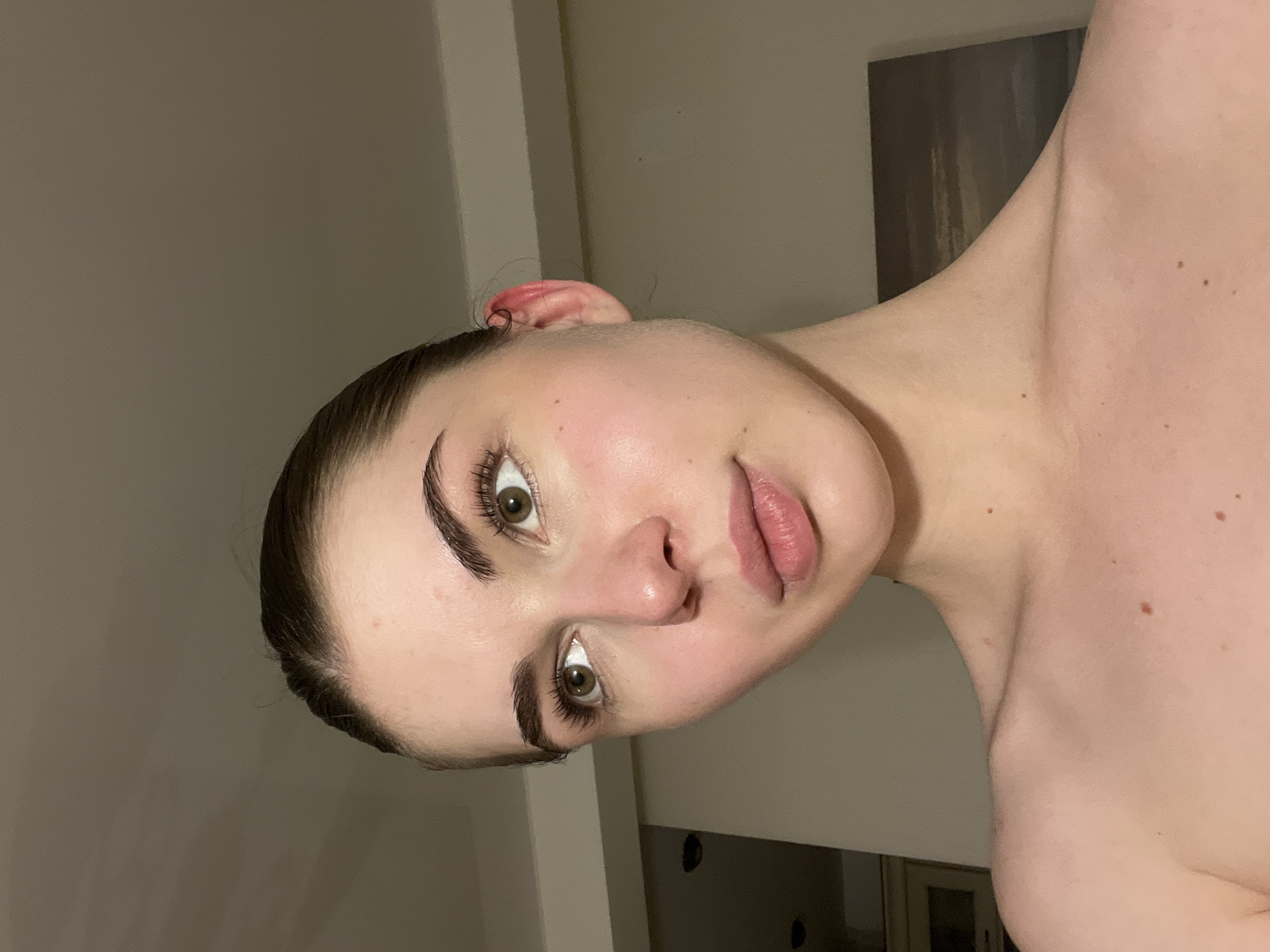 A close-up, rotated studio shot captures a woman's face and upper torso. She has fair skin, dark hair pulled back, and defined eyebrows. Her eyes are hazel, and her lips are a neutral pink. The background is a muted grey tone with vertical architectural elements, potentially wall trim or a door frame. A partially visible, darker abstract artwork hangs on the wall to the upper right. The woman's exposed shoulder and neck reveal a few small moles. The environment appears to be indoors. There is no information about the city of Saint Petersburg, Russia, in the image.