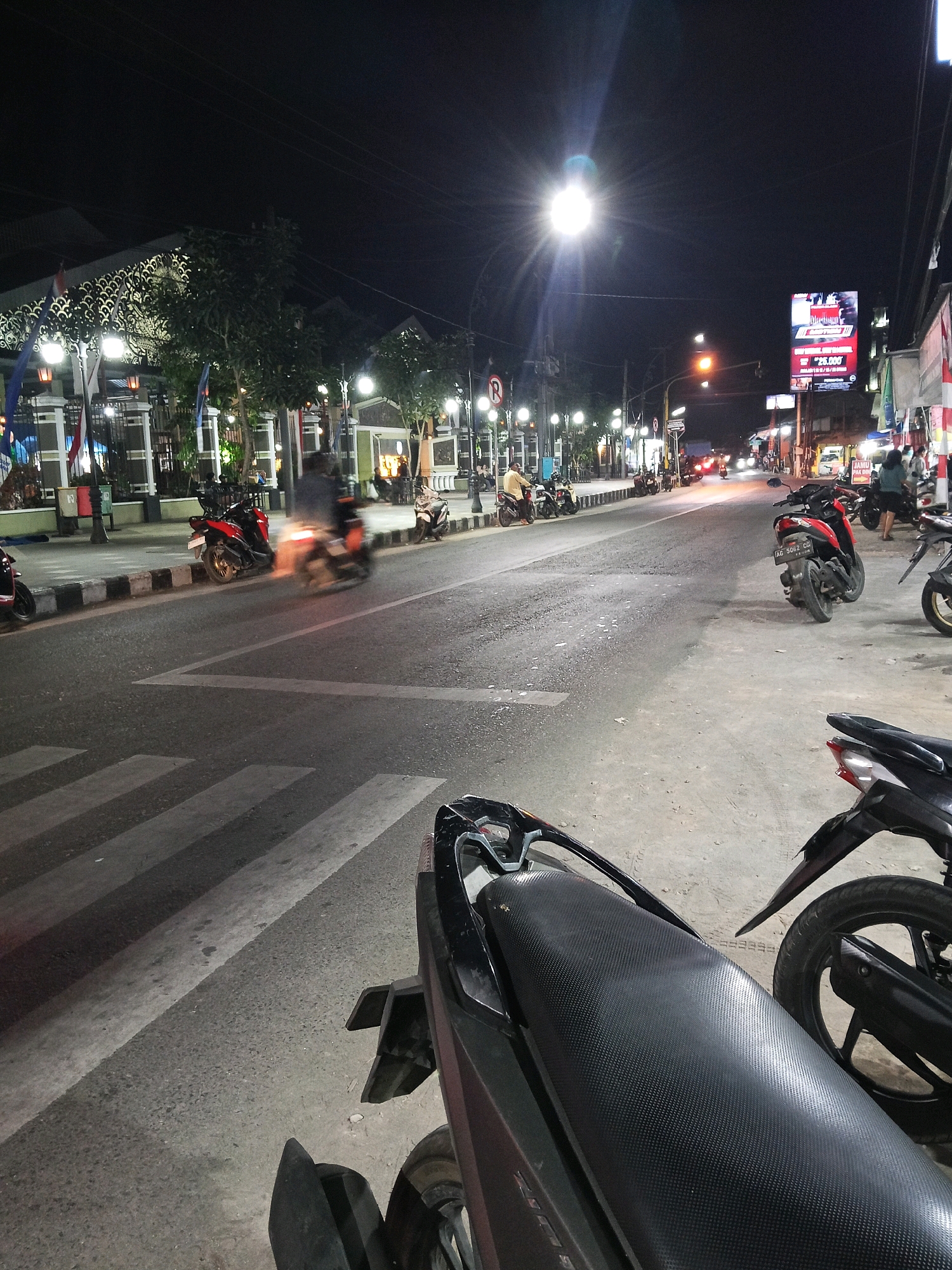 The image depicts a street scene at night in Wates, Indonesia. A busy street is visible, with several motorcycles parked along the sidewalk and others in motion. Buildings line one side of the street, featuring storefronts with lights and signage. A large billboard advertising a price of "25.000" is prominently displayed. The street is illuminated by streetlights and the headlights of vehicles. A pedestrian crossing is marked on the asphalt. The overall atmosphere suggests a commercial or social area active during the evening.