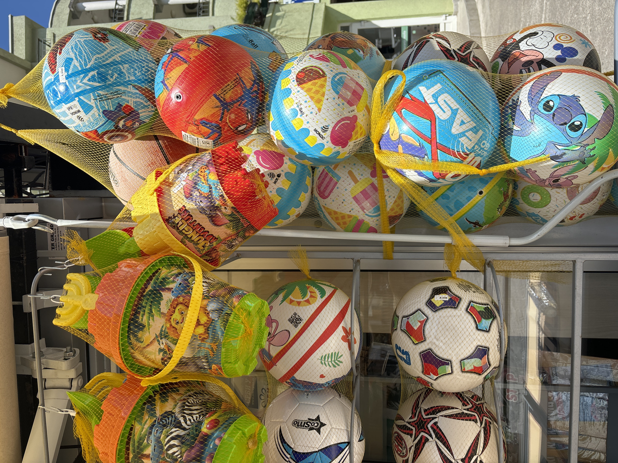 A collection of colorful beach balls and sand toys hang in mesh bags from a metal rack. The beach balls feature various designs including cartoon characters like Stitch, ice cream cones, and geometric patterns. Below the balls are hanging sets of plastic sand toys, including buckets, shovels, and rakes, also contained in mesh bags. Text and logos such as "Cosmo", "JUNGLE ADVENTURE", "FAST", "BioBall", and "MONDO" are visible on the balls and packaging. Based on the Cyrillic text and euro currency prices, this image was taken in Budva, Montenegro.