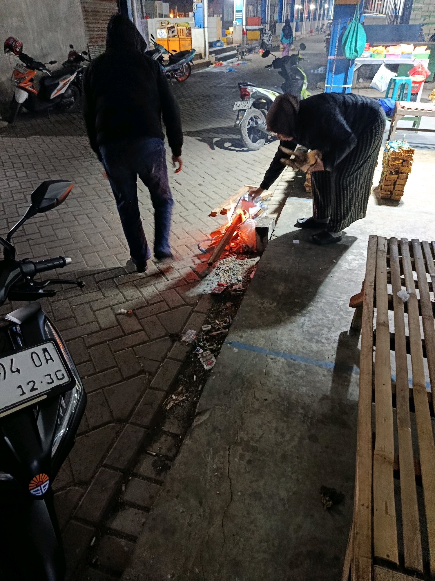 In an outdoor urban setting, potentially a market or street in Malang, Indonesia, a man wearing a black hoodie and dark pants walks away from a small bonfire. To the right, a person in a dark headscarf and striped skirt bends over the fire, tending to it with pieces of wood. In the foreground, a black scooter with a license plate reading "94 OA 12.3G" is parked. Scattered cigarette butts and debris are visible on the paved ground. In the background, several motorcycles are parked, and market stalls with goods are illuminated by artificial lighting. A wooden bench is positioned to the right of the frame.
