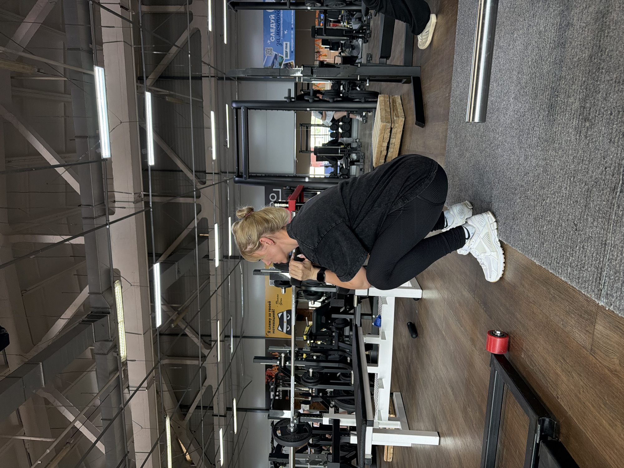 A woman is crouched in a gym in Chaikovsky, Russia, performing a weight training exercise on a seated row machine. She is wearing black leggings, a dark gray t-shirt, and white athletic shoes. Her blonde hair is pulled back. She is holding the handles of the machine with both hands, appearing to engage her back muscles. The gym is well-equipped with various weight machines, and the ceiling has exposed metal beams and fluorescent lights. A sign with Russian text is visible on the machine. The floor is a combination of wood and gray carpet.