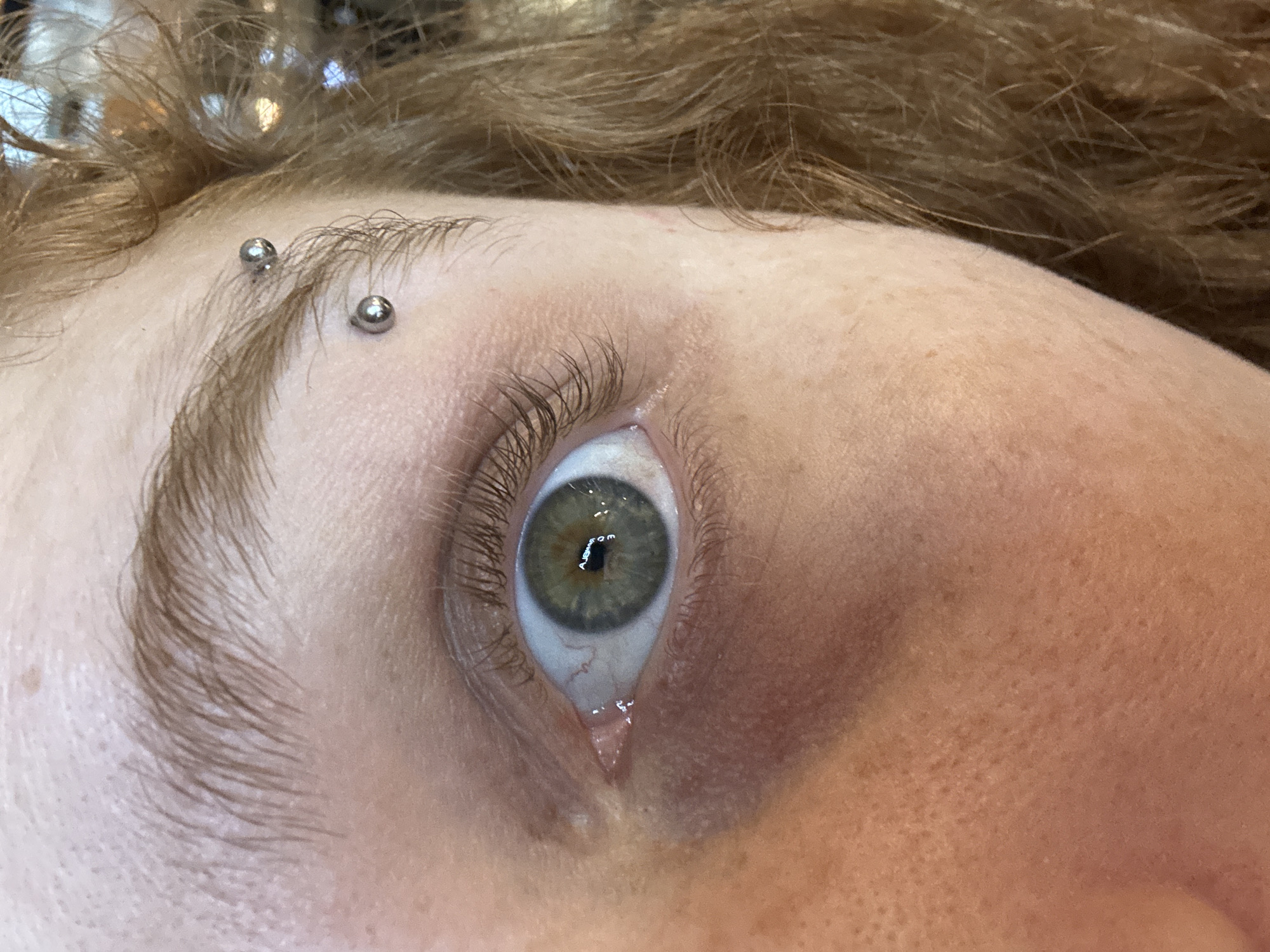A close-up shot focuses on a person's face, highlighting their left eye and the surrounding area. The eye is open, exhibiting a green iris and a clear reflection. A single tear is visible, trailing down from the lower eyelid. Above the eye, the eyebrow is pierced with two small, silver-colored studs. The skin around the eye shows some discoloration, possibly indicating tiredness or bruising. The person has light brown hair. The image does not provide information to suggest the location is in Buenos Aires, Argentina.