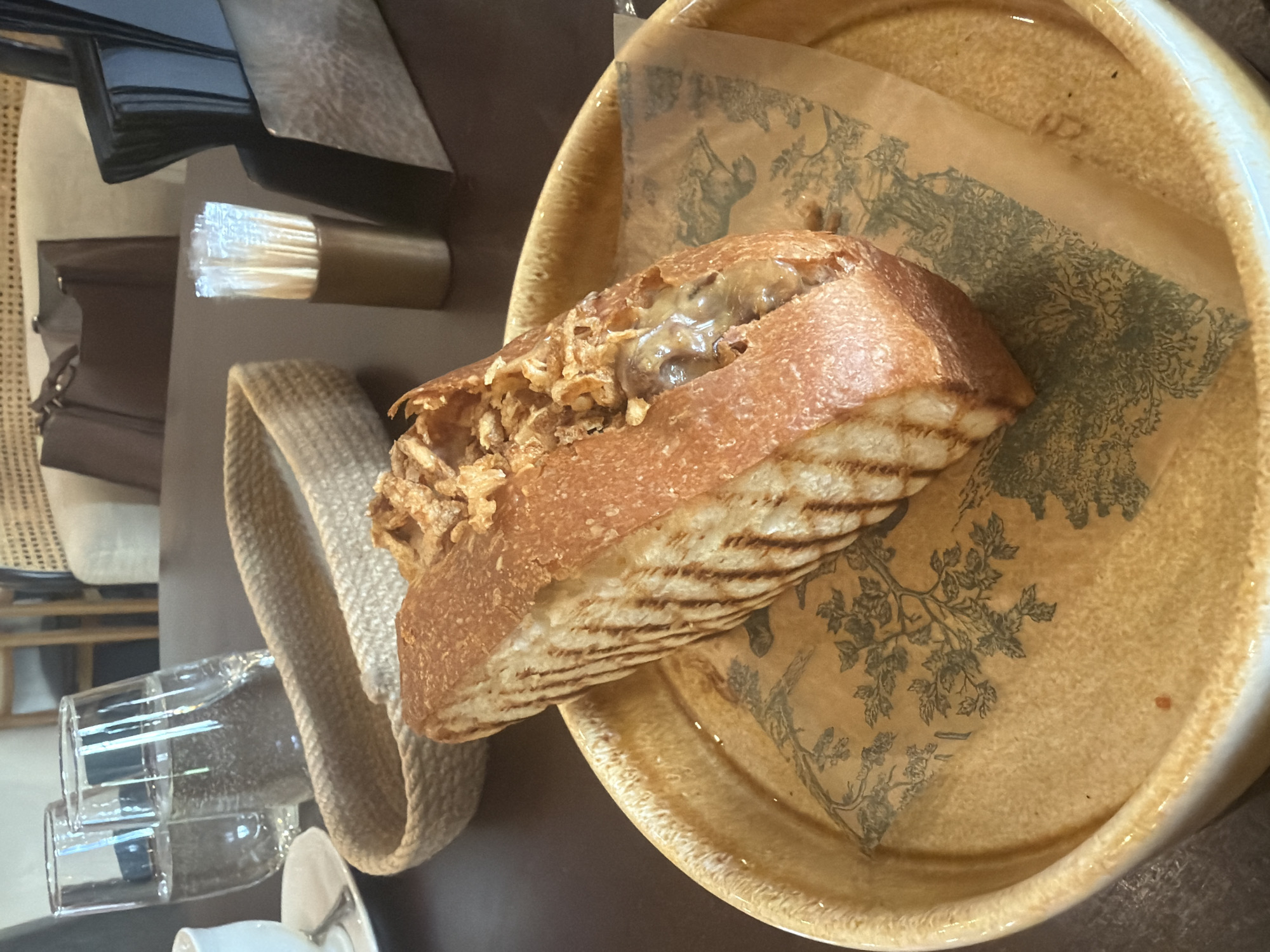 The image shows a half of a grilled bread sandwich, likely a baguette or similar bread, topped with fried onions and a creamy sauce, resting on a decorative paper liner in a shallow, wide-rimmed ceramic bowl. Beside the bowl is a woven basket and behind the bowl, closer to the camera, are a few empty glasses and a white plate and saucer, possibly a coffee set. Further in the background, there's a container holding what appears to be toothpicks, a brown purse, and a black patent leather bag.
While I don't have specific information about the exact location or the city of Krasnoyarsk, Russia, based on the image, the setting appears to be inside a café or restaurant. The composition suggests a casual, somewhat upscale dining experience. Without additional data, it's impossible to ascertain specific information such as the restaurant's name or exact locale in Krasnoyarsk.